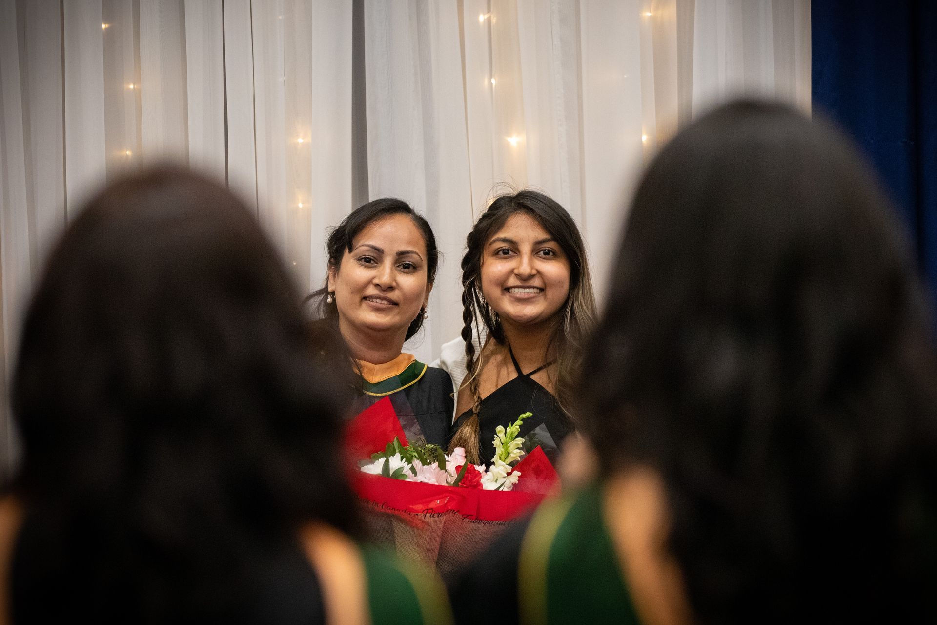 Two women stand in front of white drapes, holding flowers and smiling at an event.