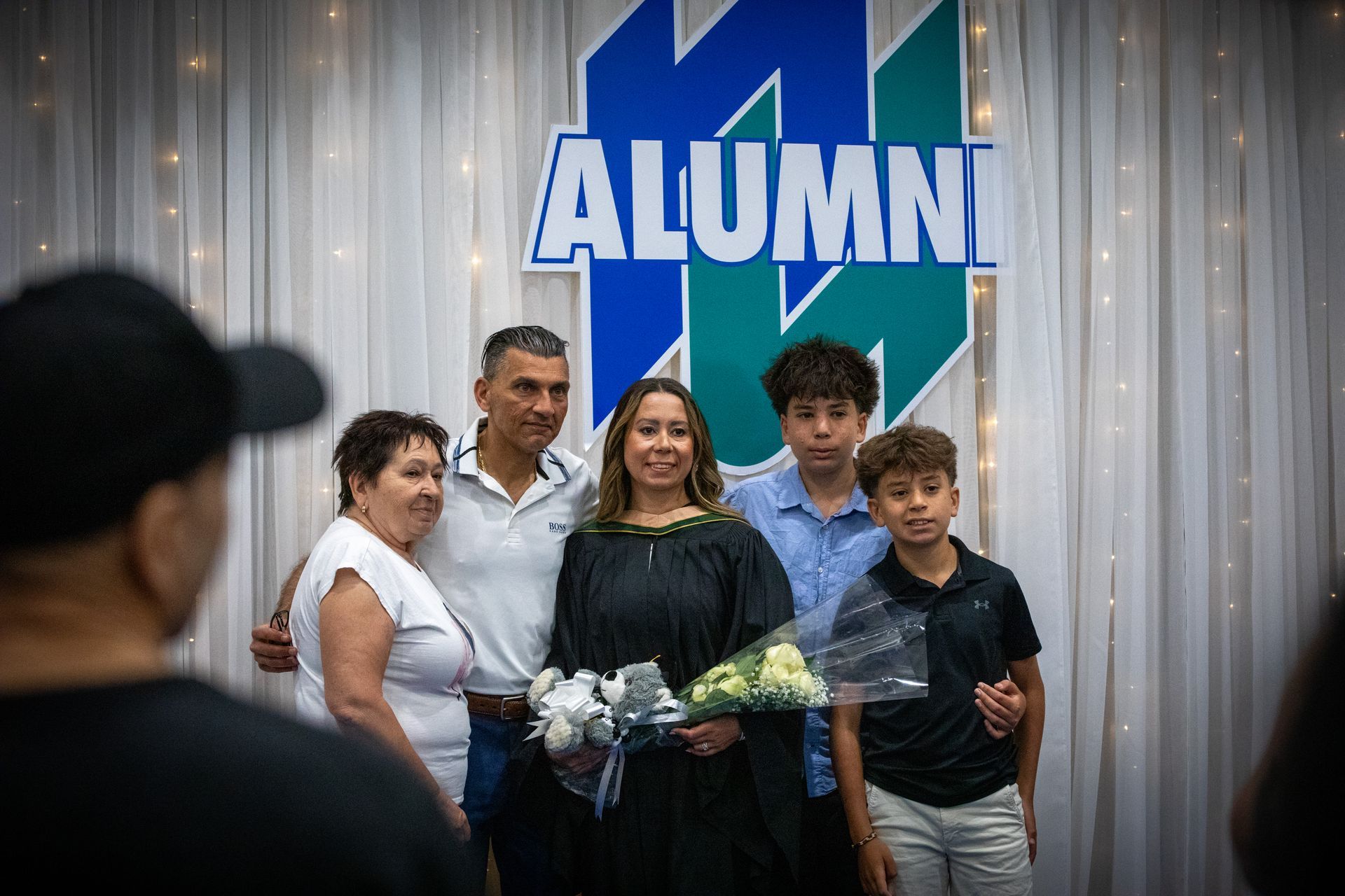 Five people pose before a blue-and-green “ALUMNI” backdrop, holding flowers at a celebration.