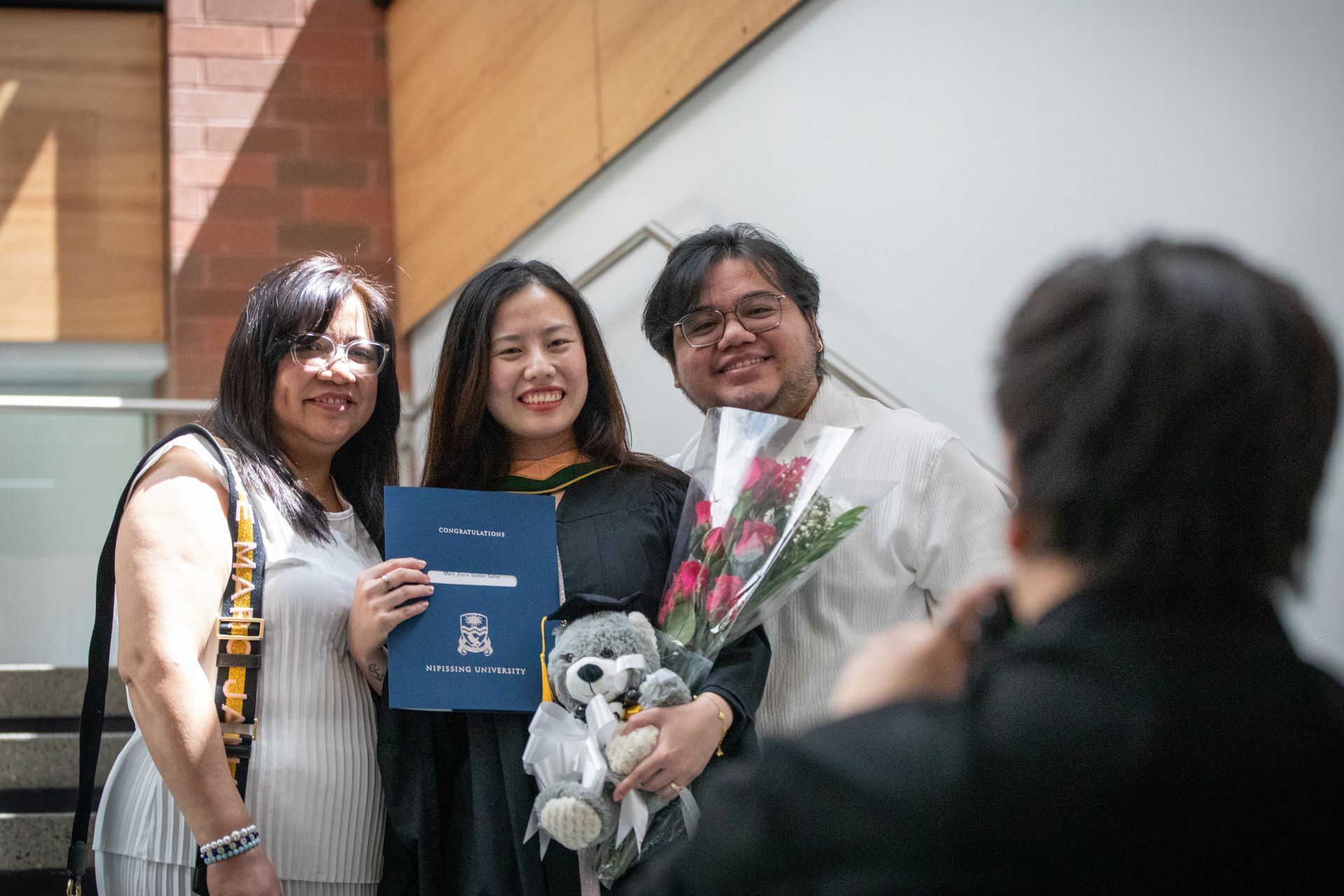 Three people pose with a blue certificate and flowers at an indoor ceremony