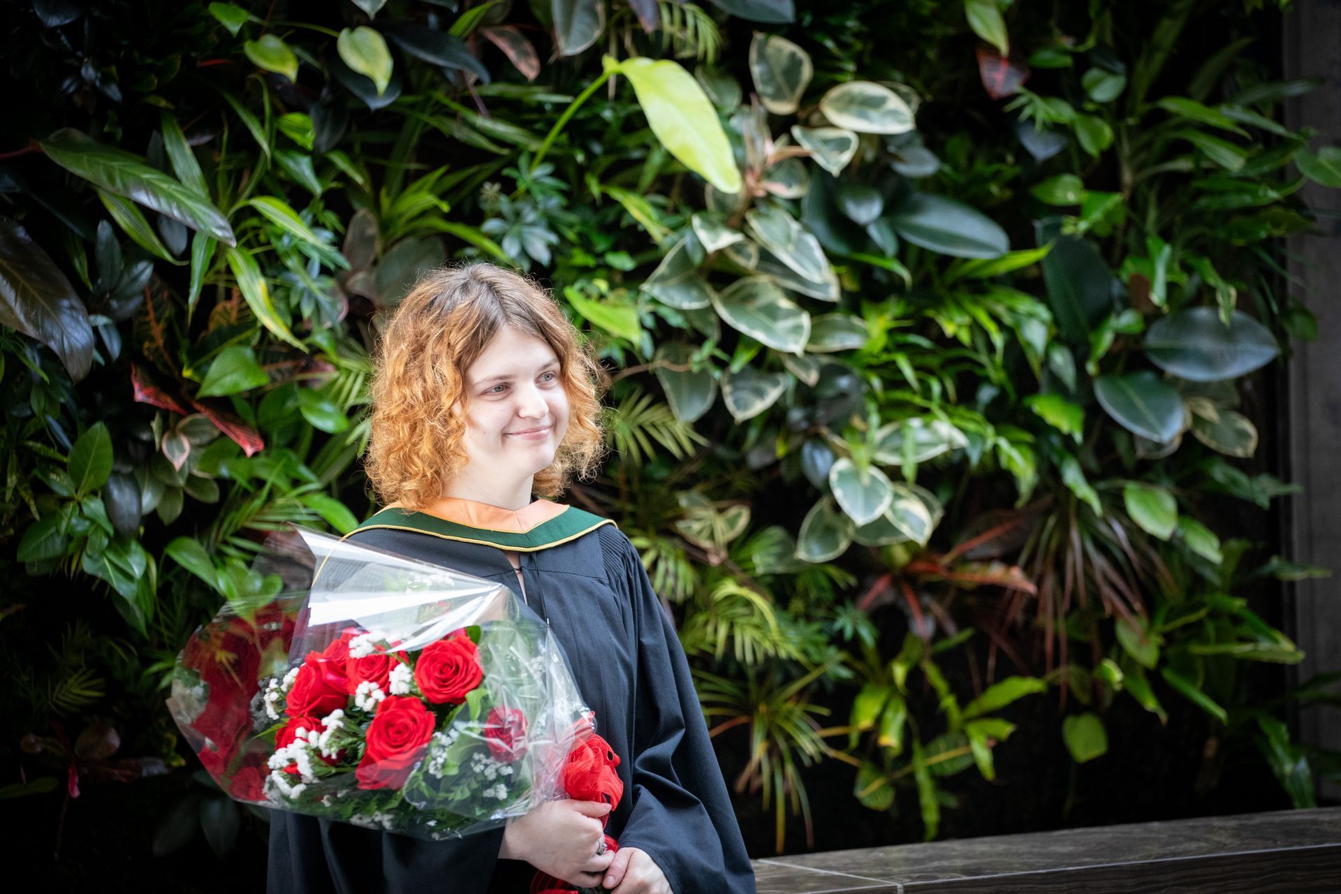 Person in a graduation gown holding a bouquet of red roses in front of lush green foliage