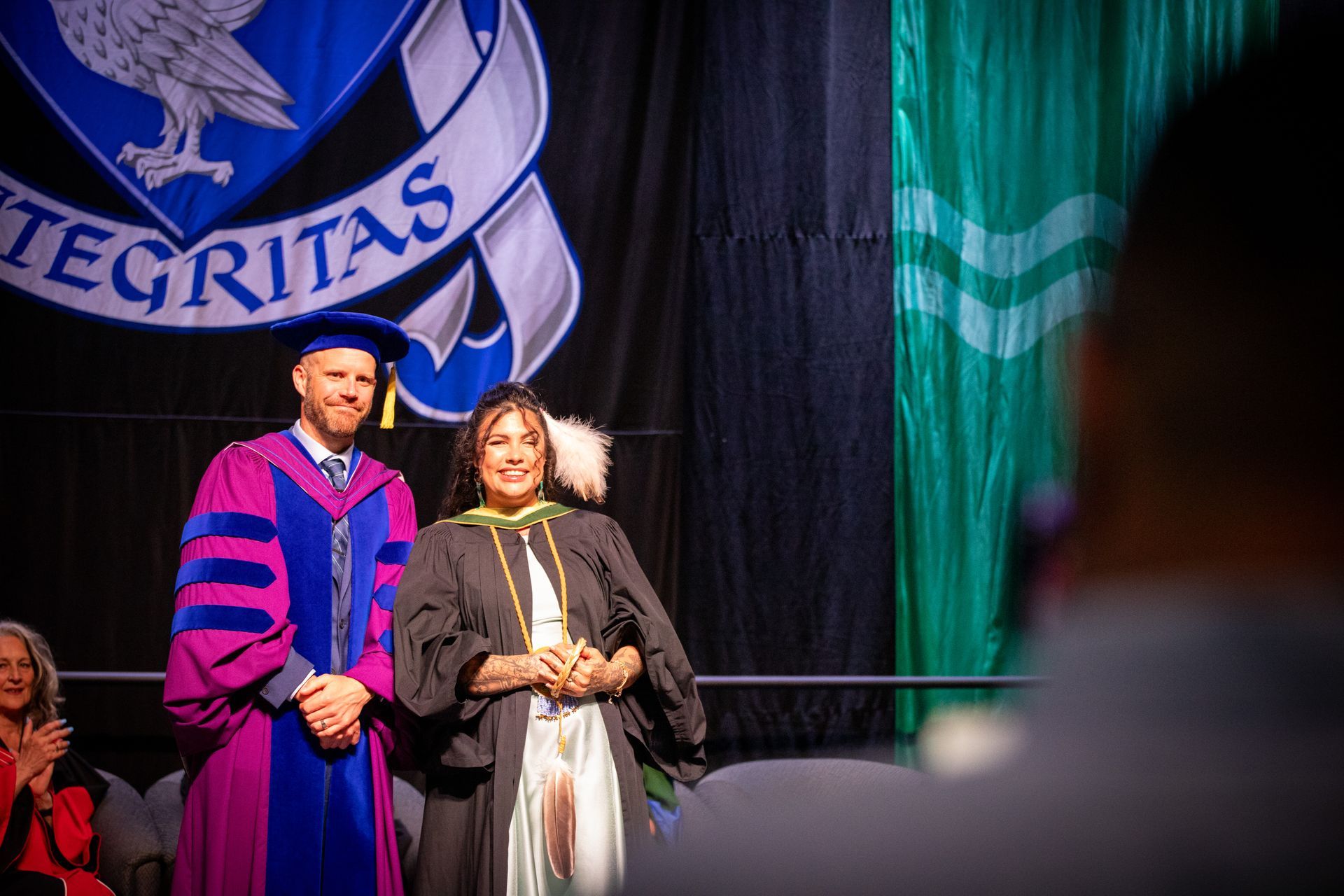 Two people in academic regalia onstage, standing before a large banner and green drapes.