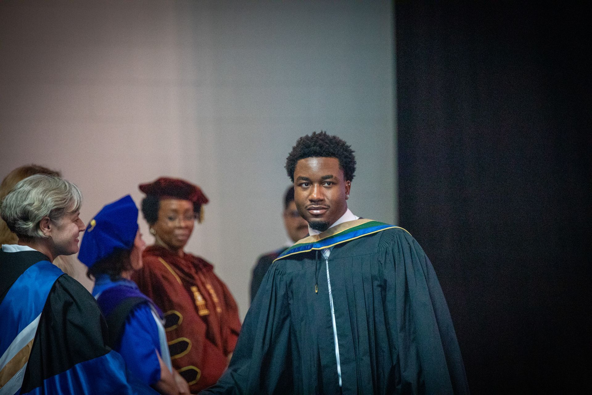 Graduate in black gown and blue hood stands before seated faculty at a commencement ceremony.