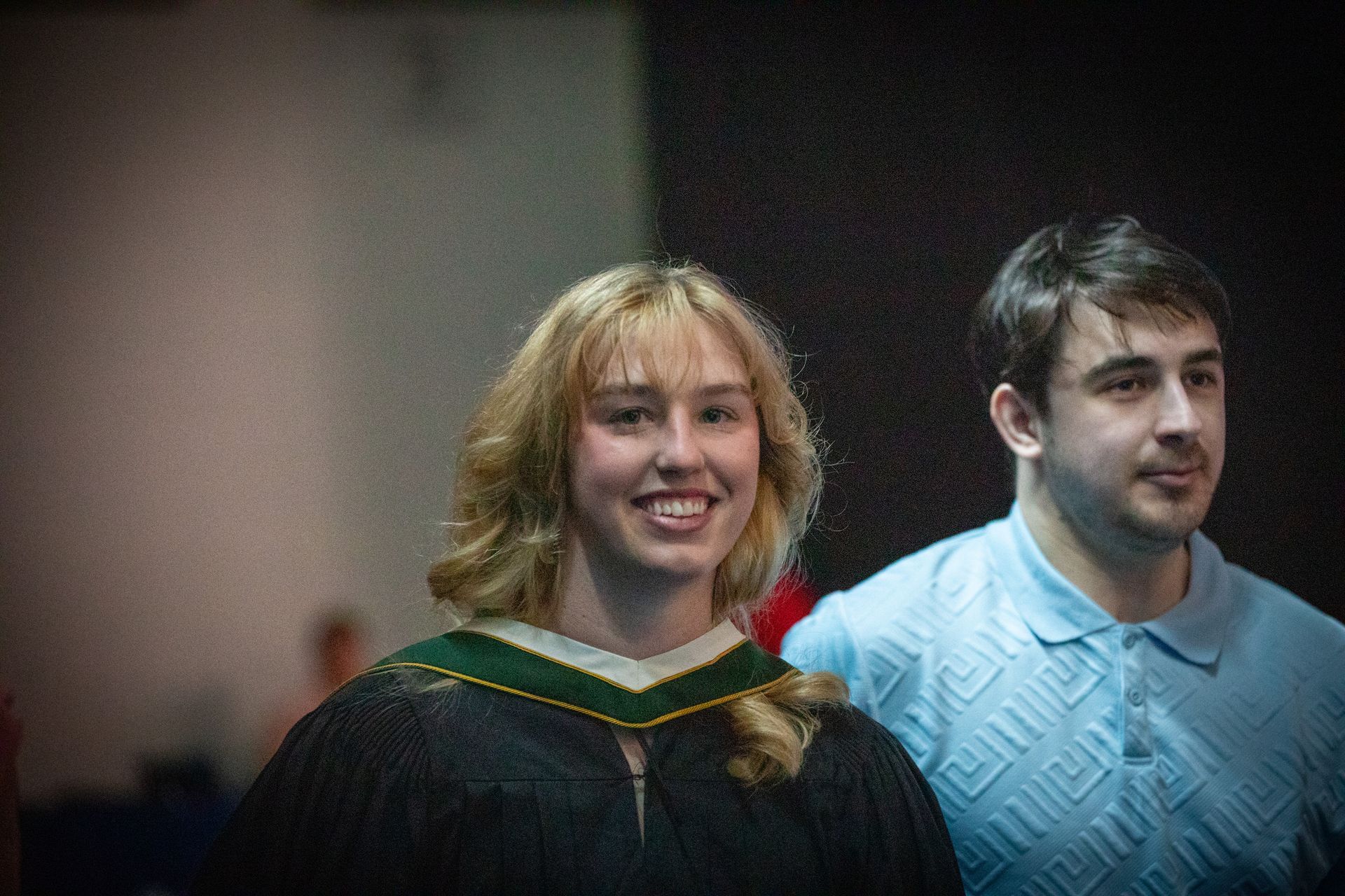 Two people smiling indoors, one in a black gown with a green-trimmed collar and one in a light blue sweater.