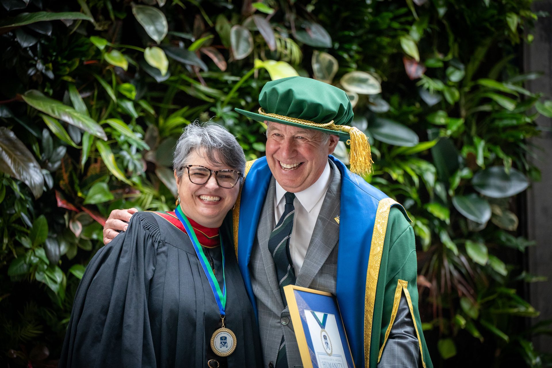 Two smiling people in caps and gowns pose together in front of greenery