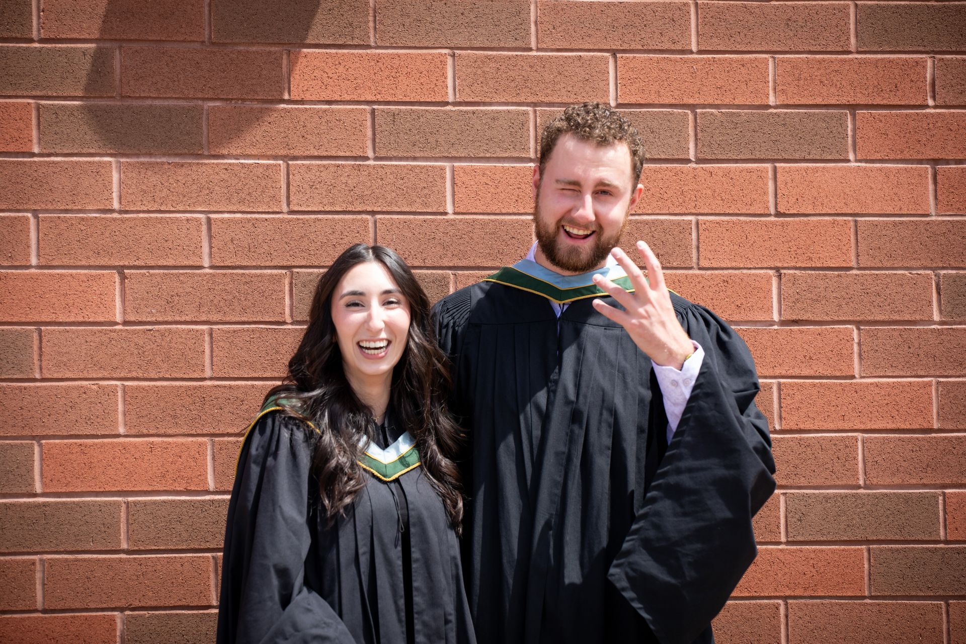 Two graduates in black gowns smiling in front of a brick wall, one holding up a hand sign.