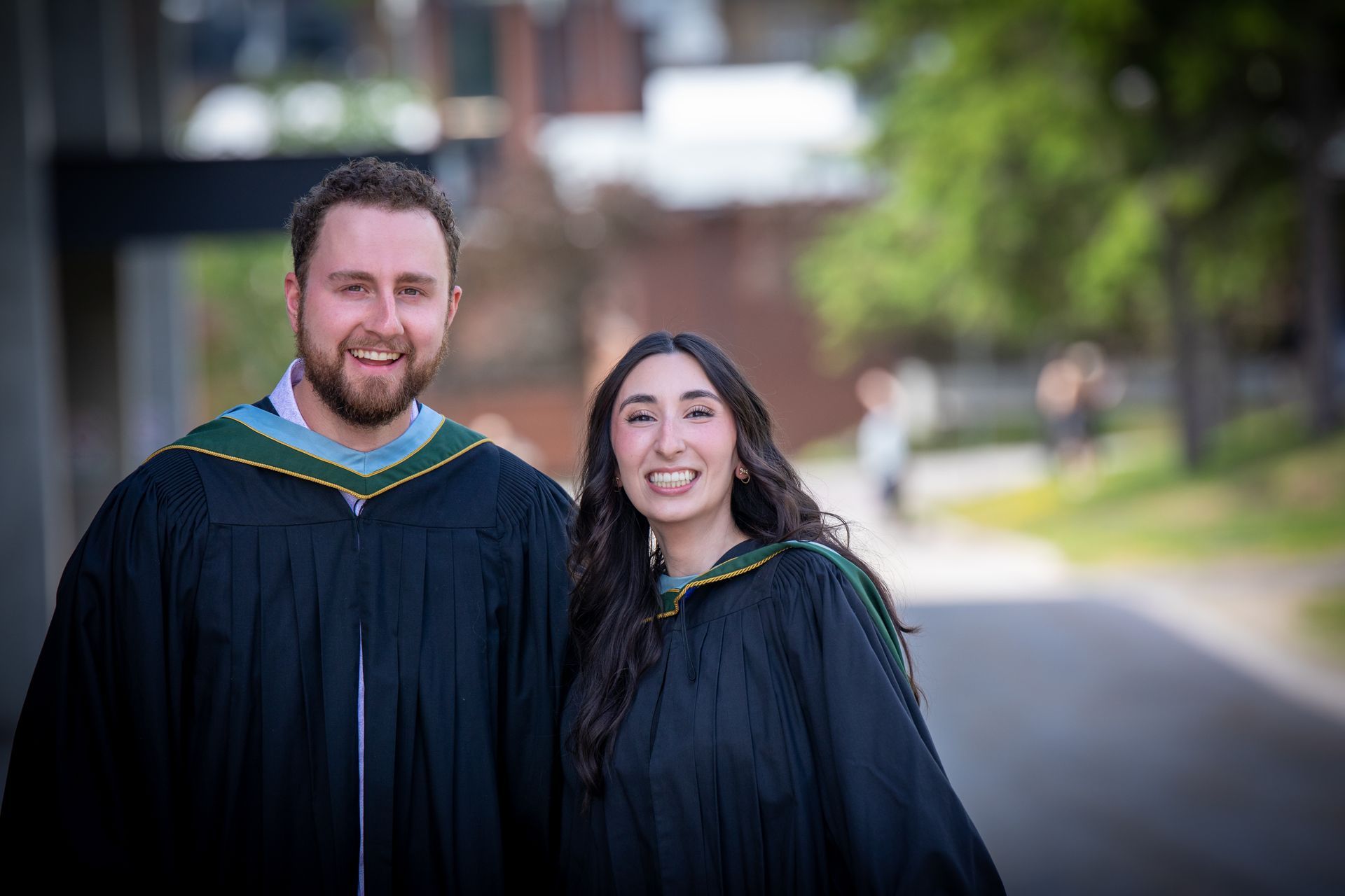 Two graduates in black gowns and green-trimmed hoods smiling outdoors on a campus walkway