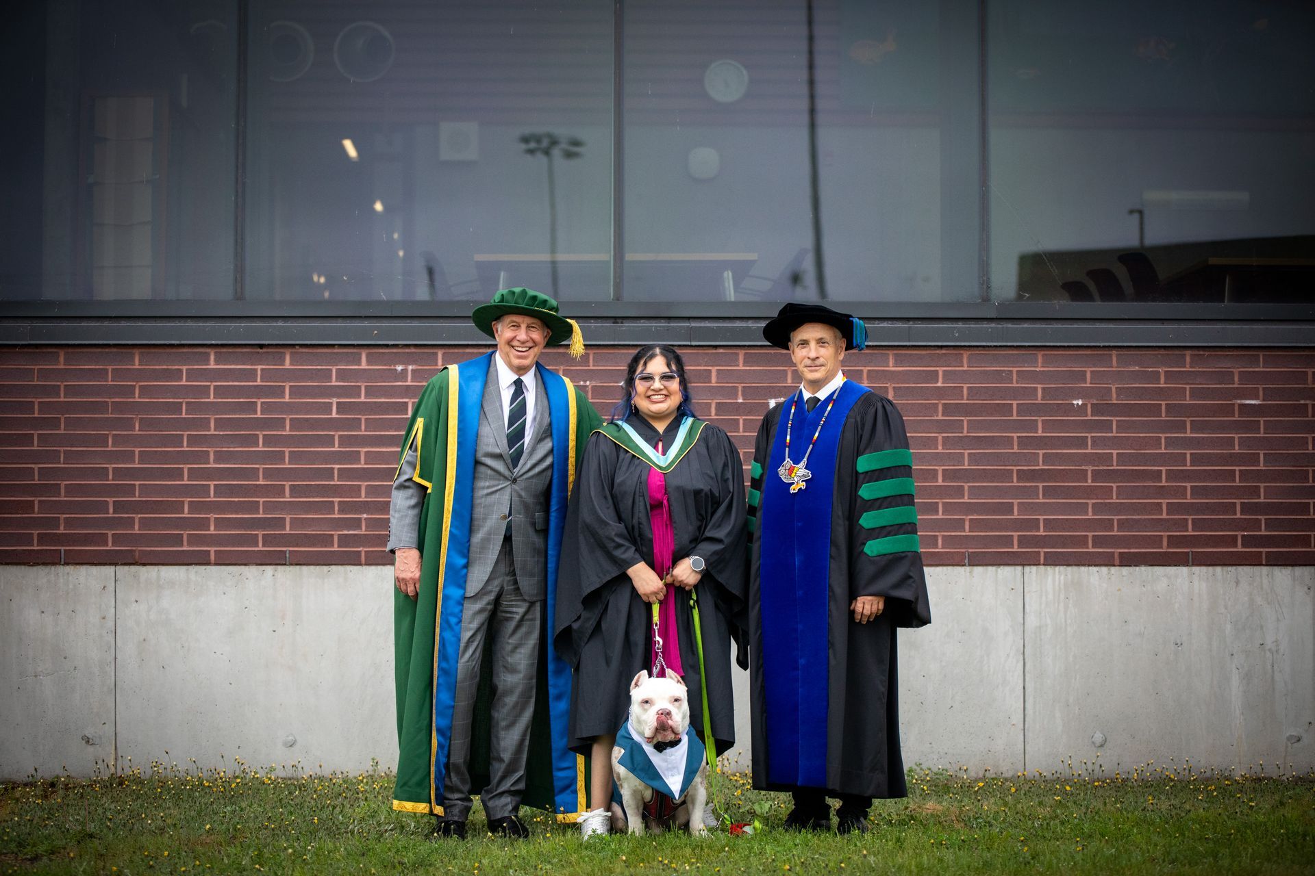 Three people in gowns pose with a dog in front of a brick building.