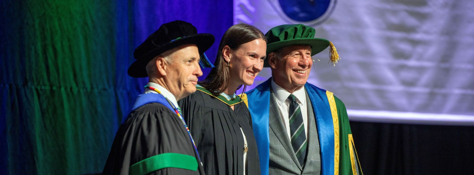 Three people in academic robes and caps standing on stage, smiling under purple lighting