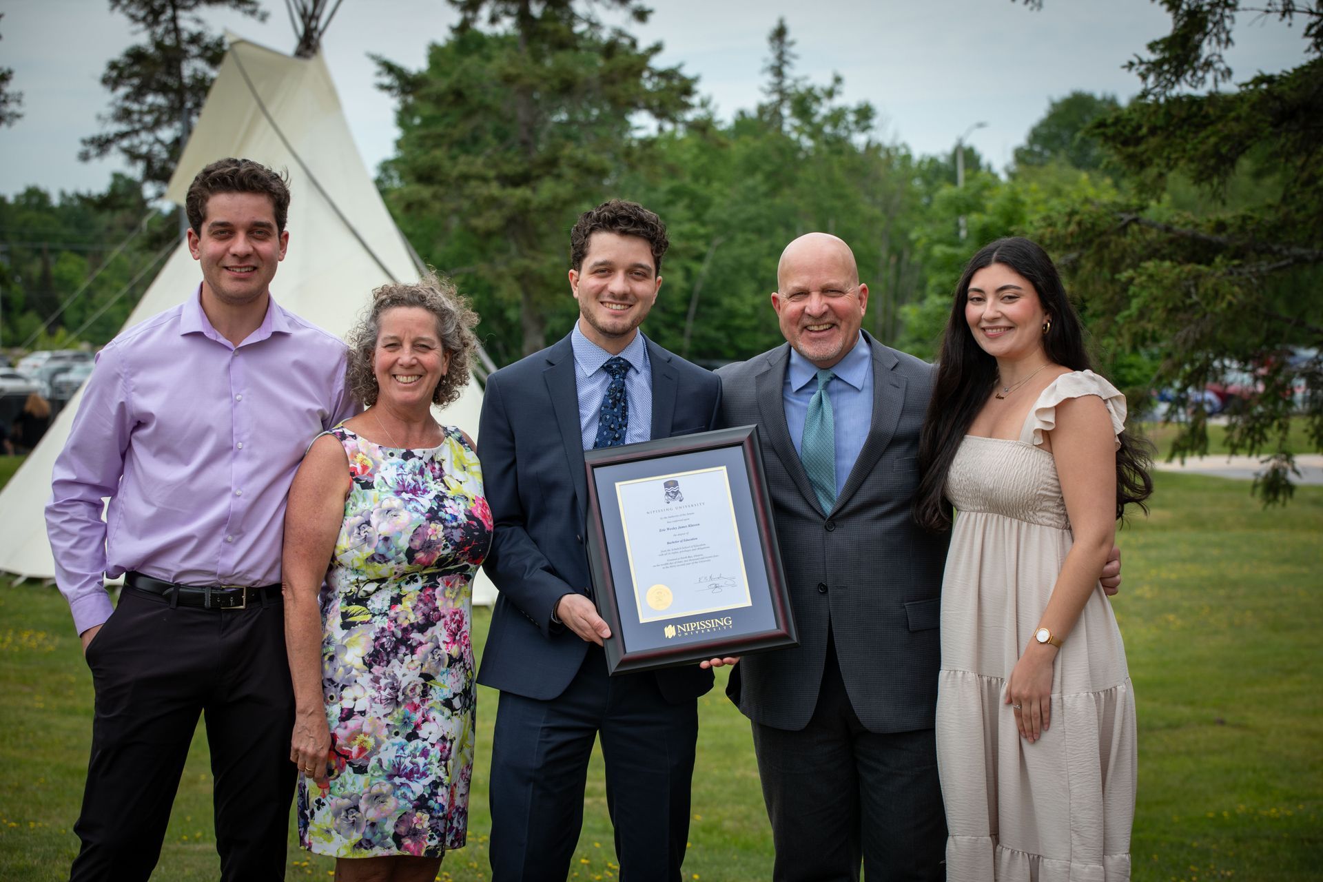 Five people posing outdoors, one holding a framed certificate, with a teepee in the background.