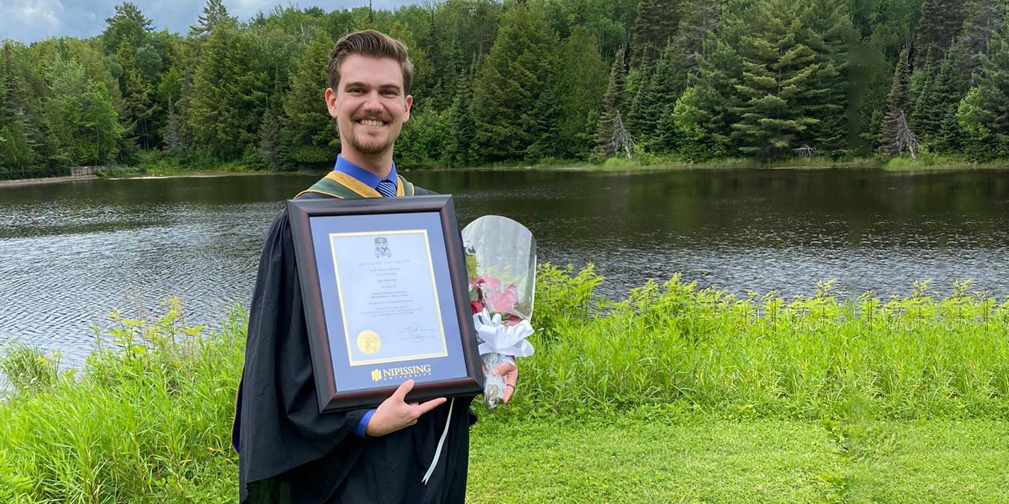 Person in graduation gown holds a framed certificate and bouquet by a lake with trees in the background
