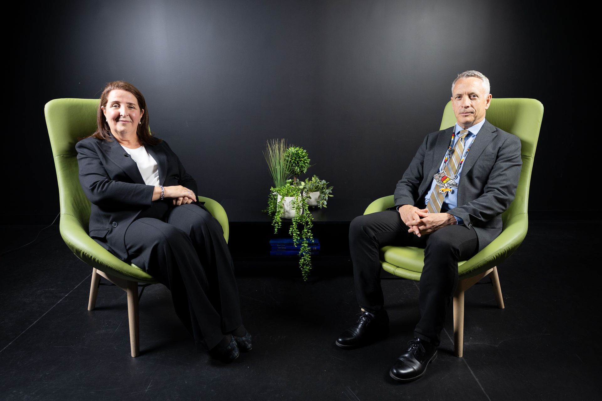 Two seated people in green chairs with a small plant between them on a dark studio set