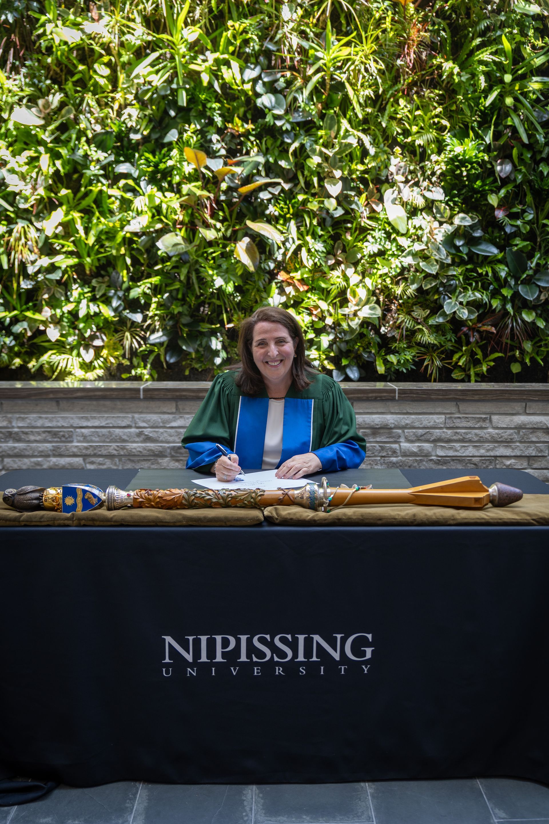 Woman seated behind a Nipissing University table outdoors, with greenery in the background.