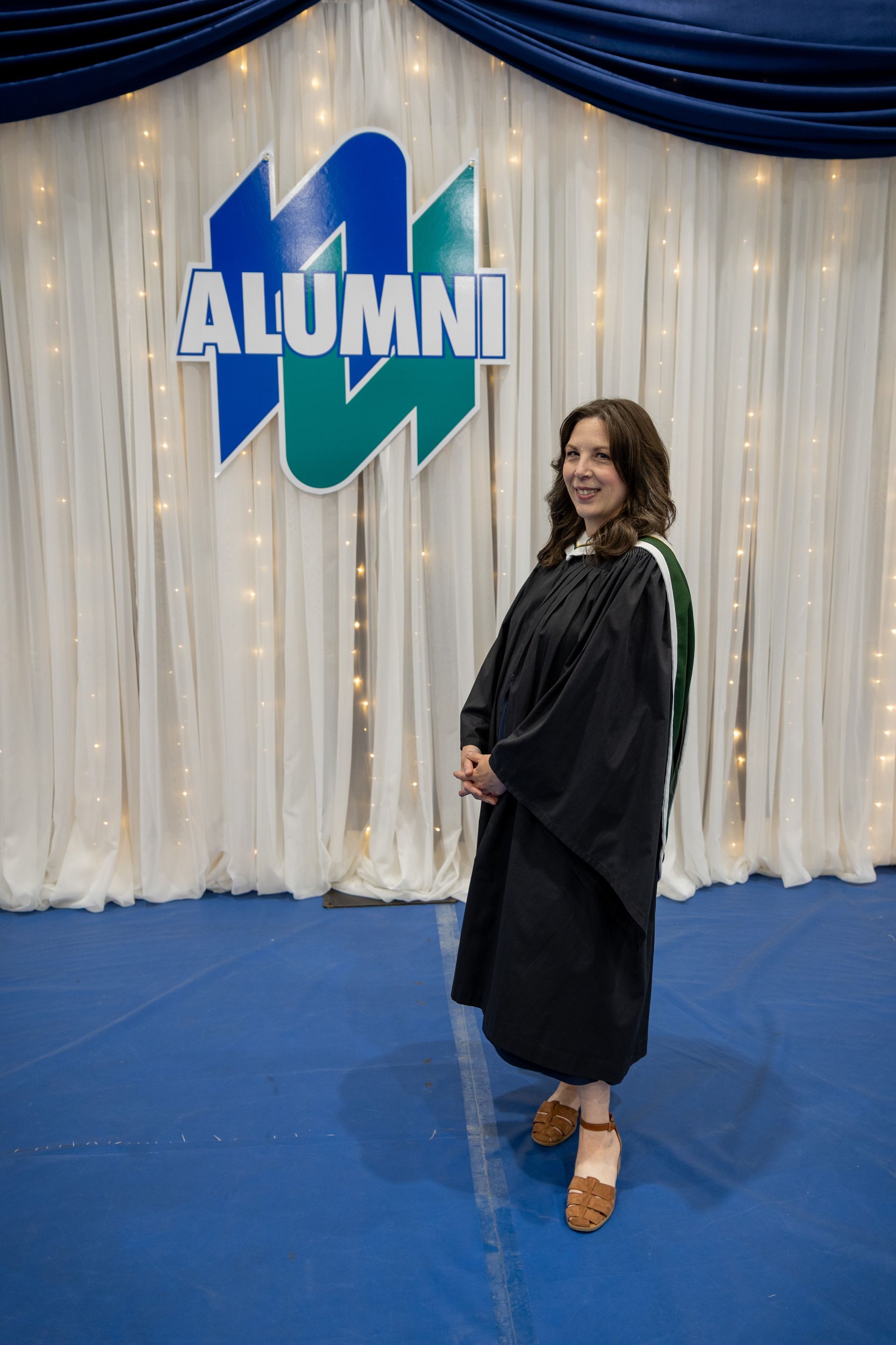 Woman in black graduation gown posing on a blue carpet before an “ALUMNI” backdrop.