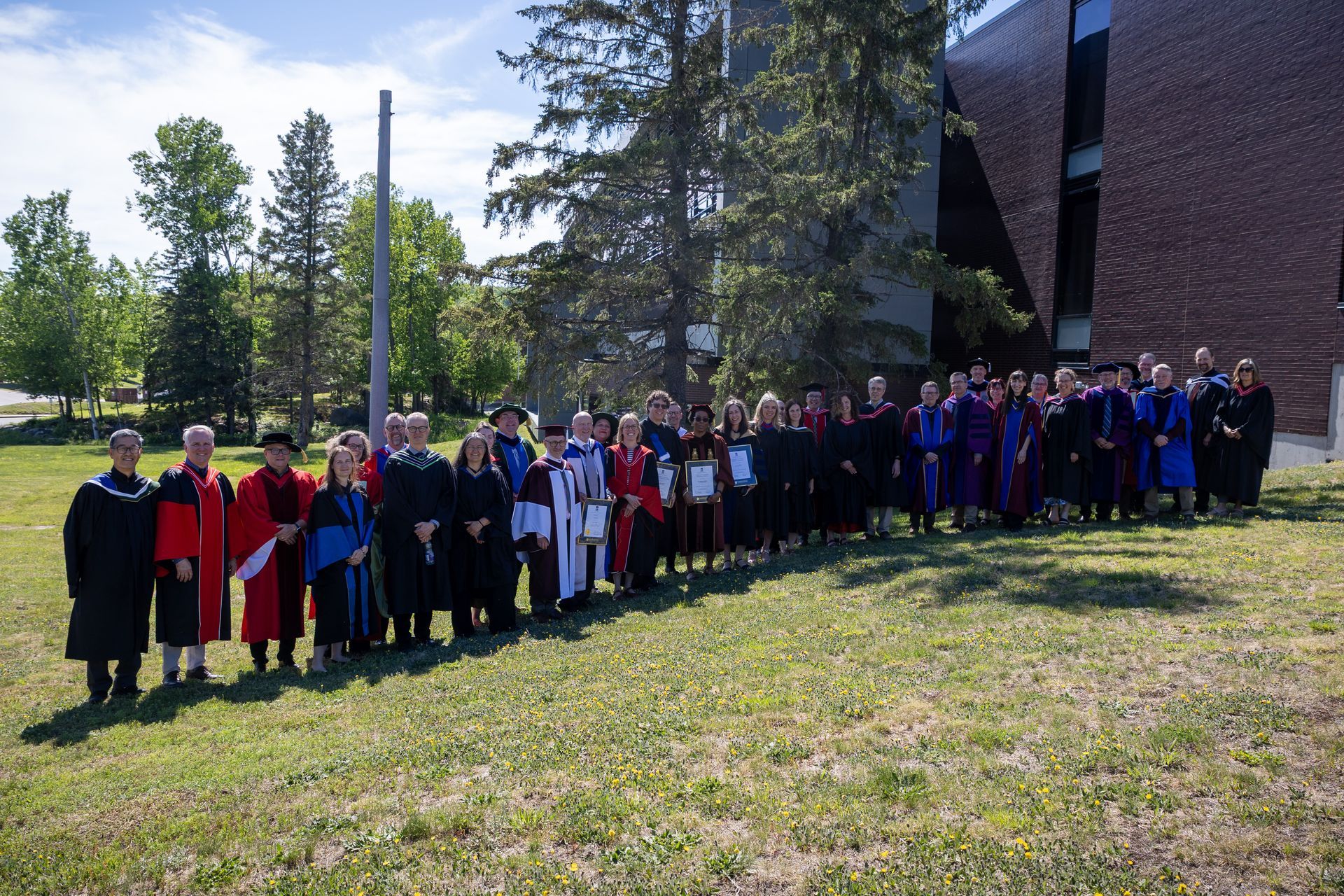 Group of people in graduation robes posing outdoors beside a brick building and trees