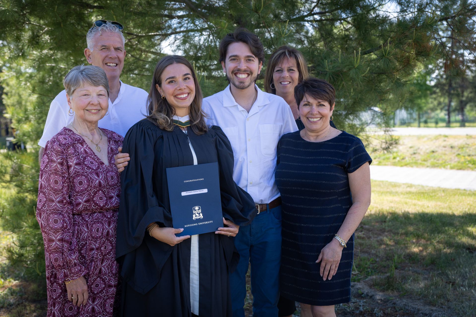 Six people standing outdoors, smiling with a graduate holding a diploma cover.