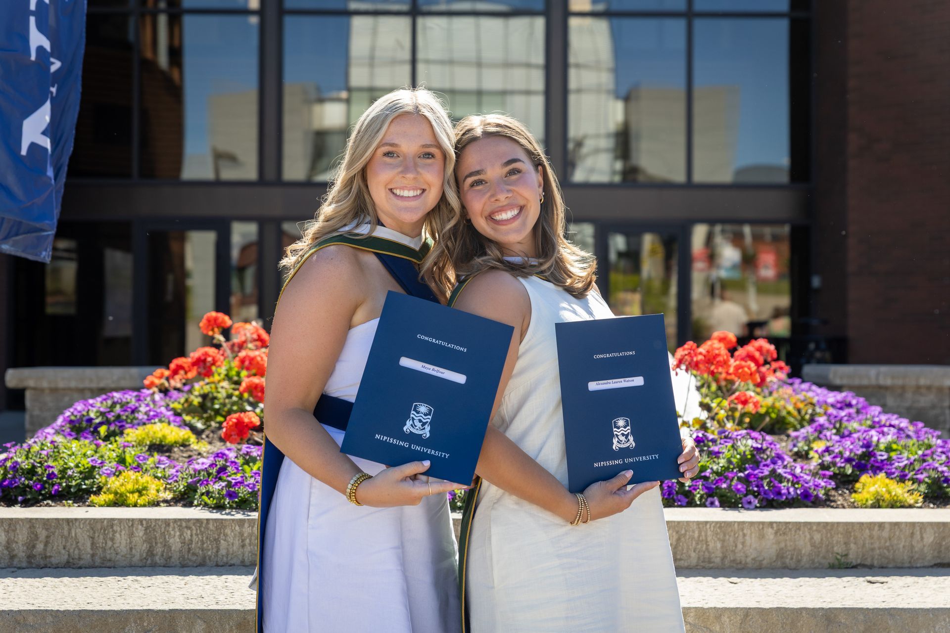 Two smiling graduates holding blue diplomas in front of a building with colorful flowers