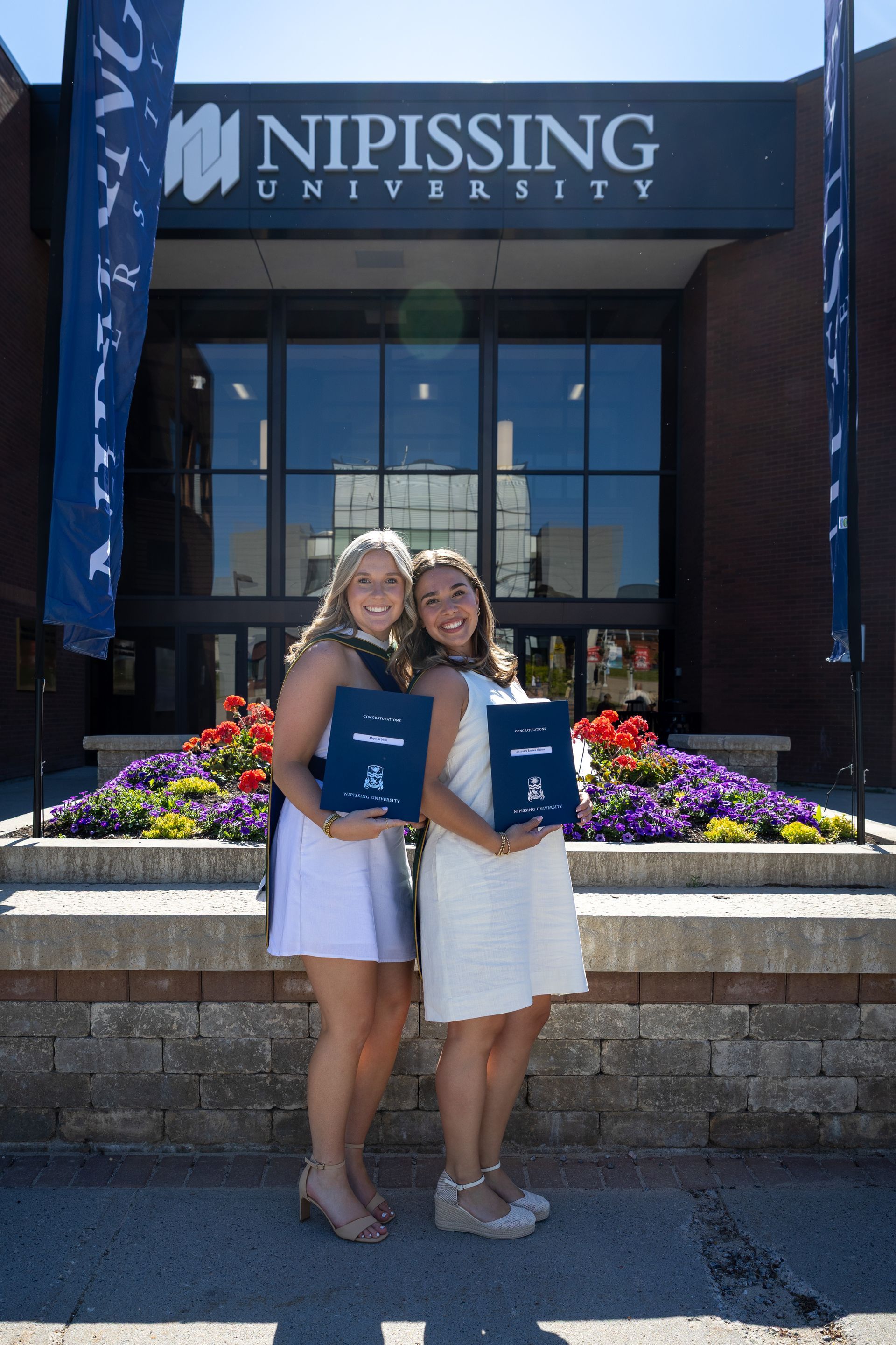 Two graduates posing outside Nipissing University, holding diplomas