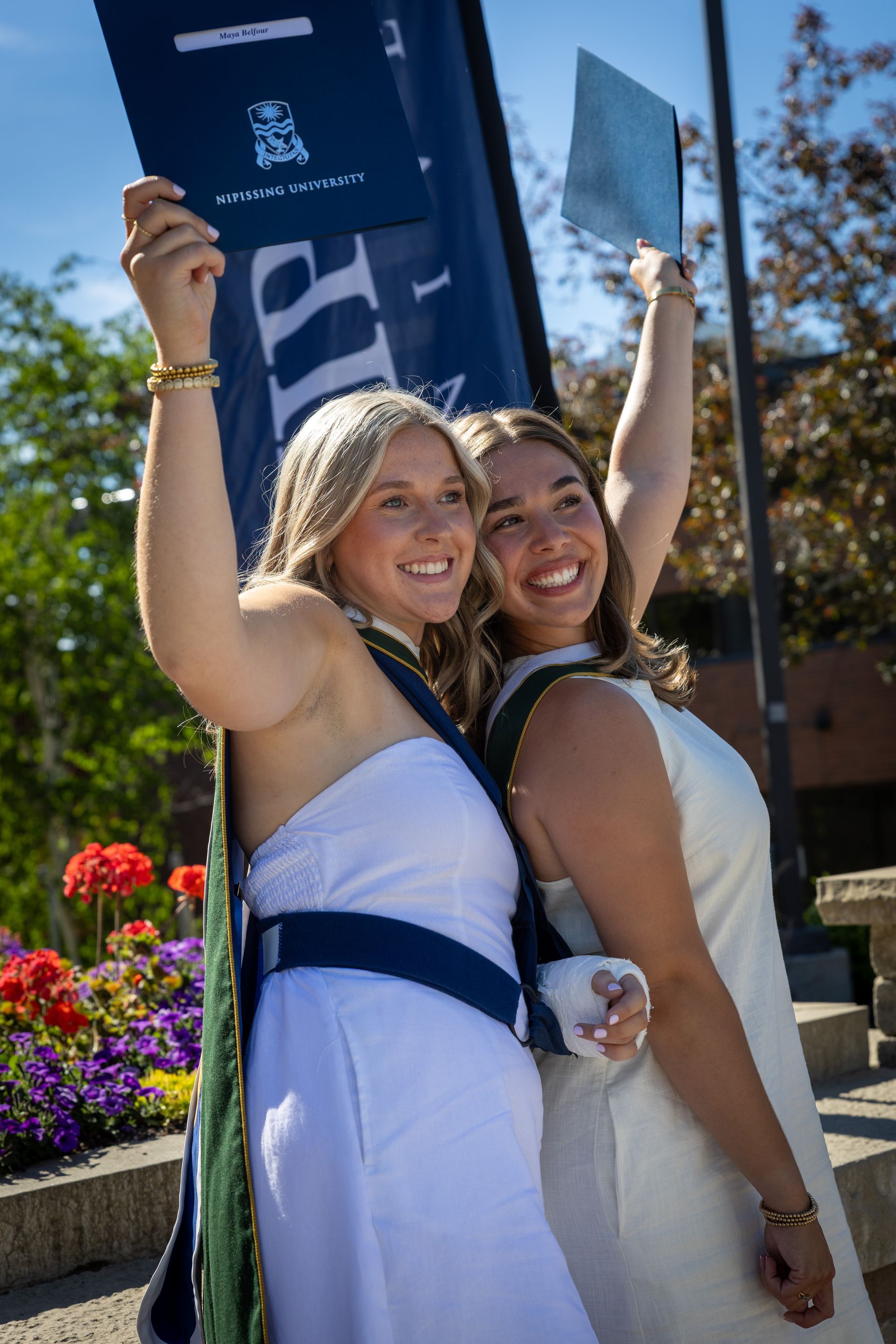 Two smiling people pose outdoors, holding up a banner near flowers and a building.