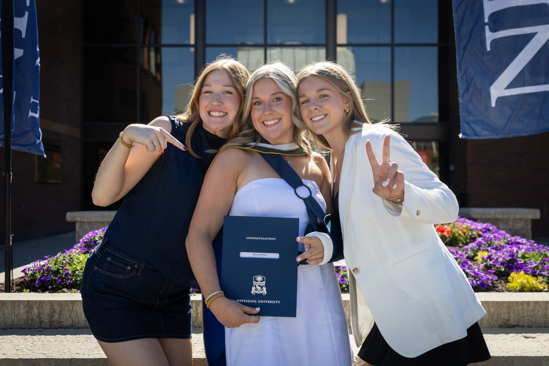 Three smiling people posing outdoors, one holding a diploma in front of a building.