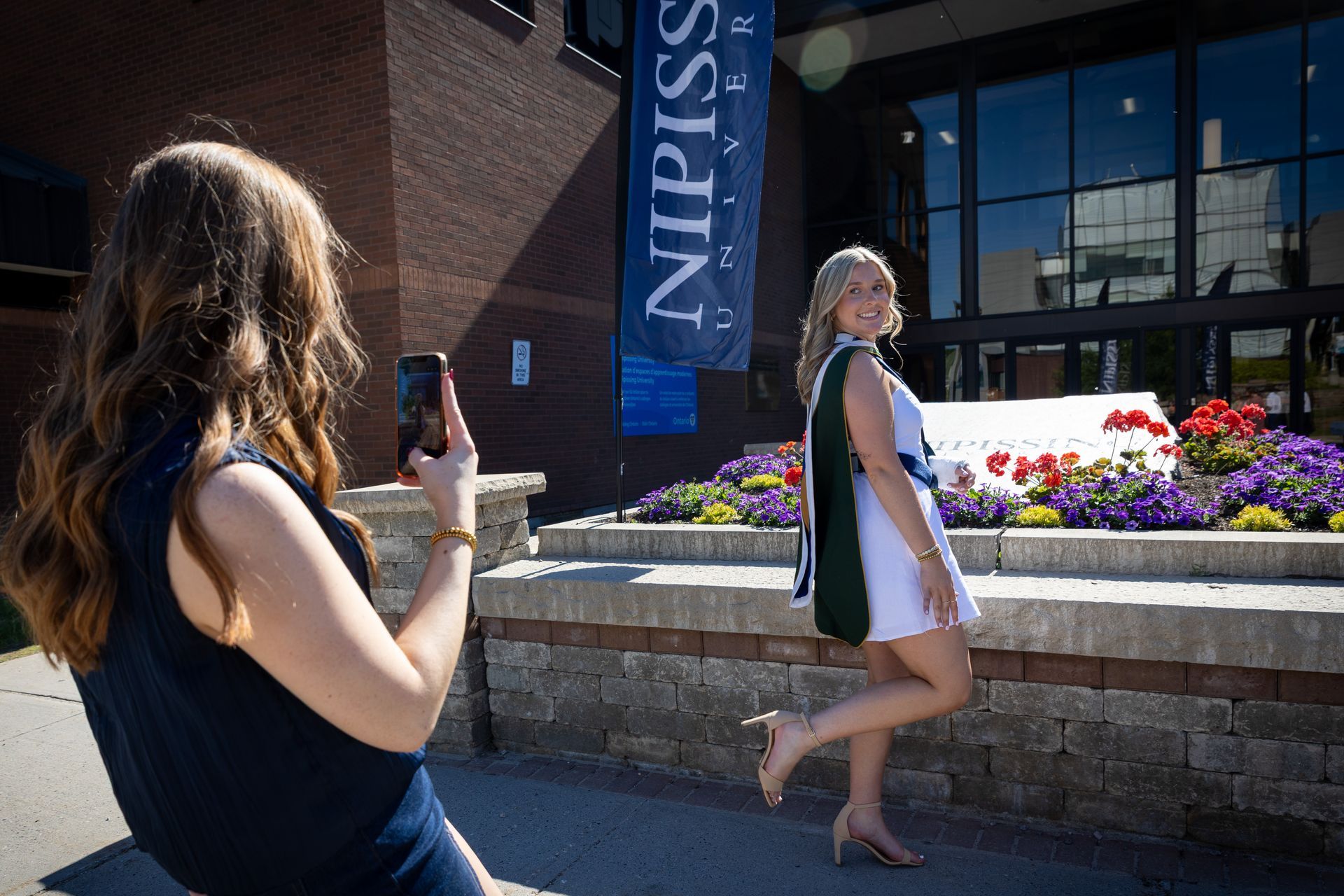 Woman posing outdoors while another person photographs her near a flower bed and building entrance