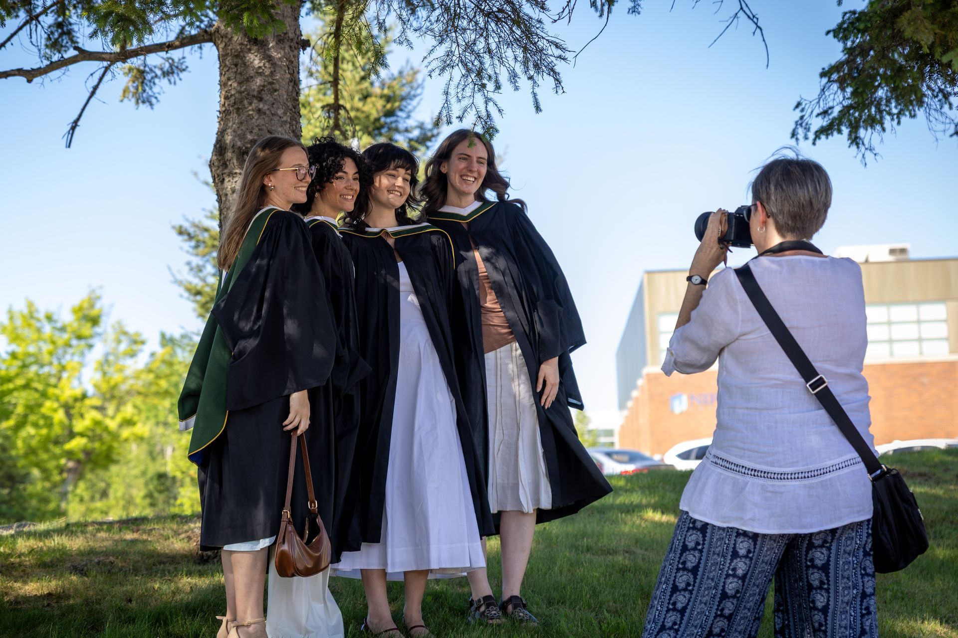 Four people in black robes pose outdoors while a photographer takes their picture.