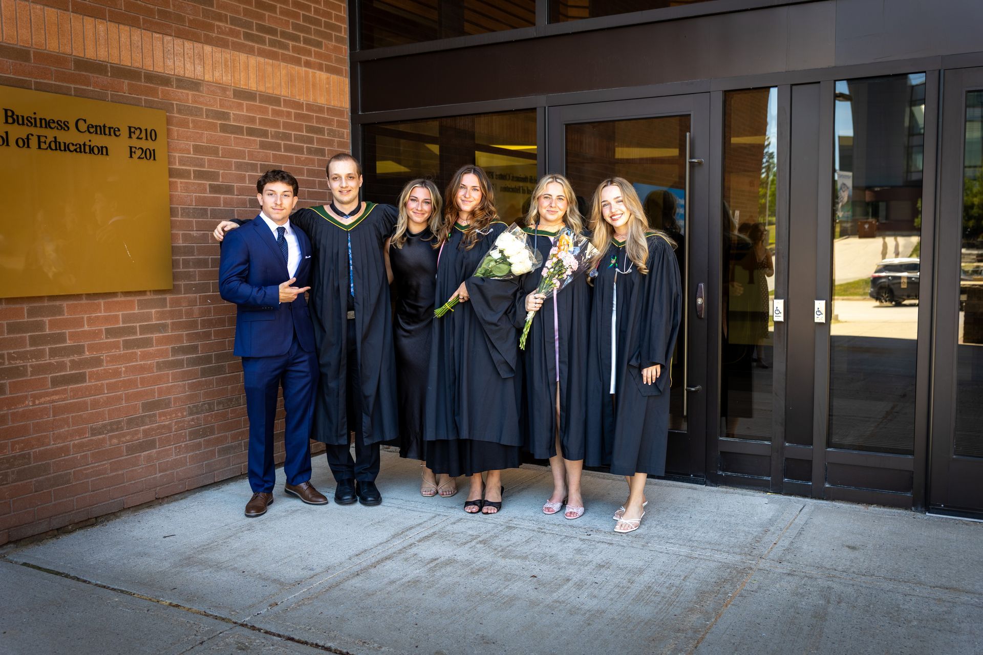 Six graduates in gowns pose outside a brick building after a ceremony.