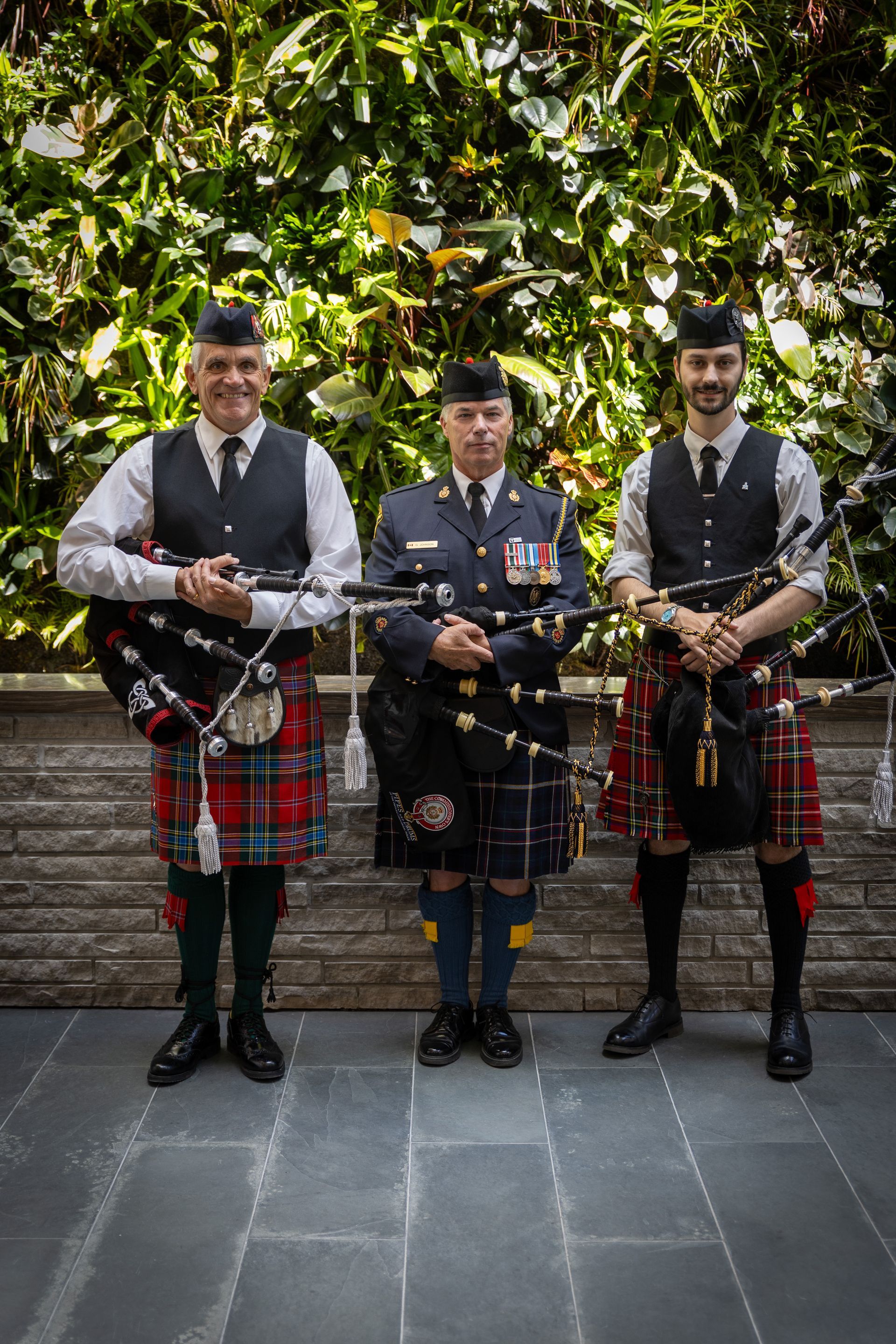 Three bagpipers in kilts standing outdoors against a leafy wall