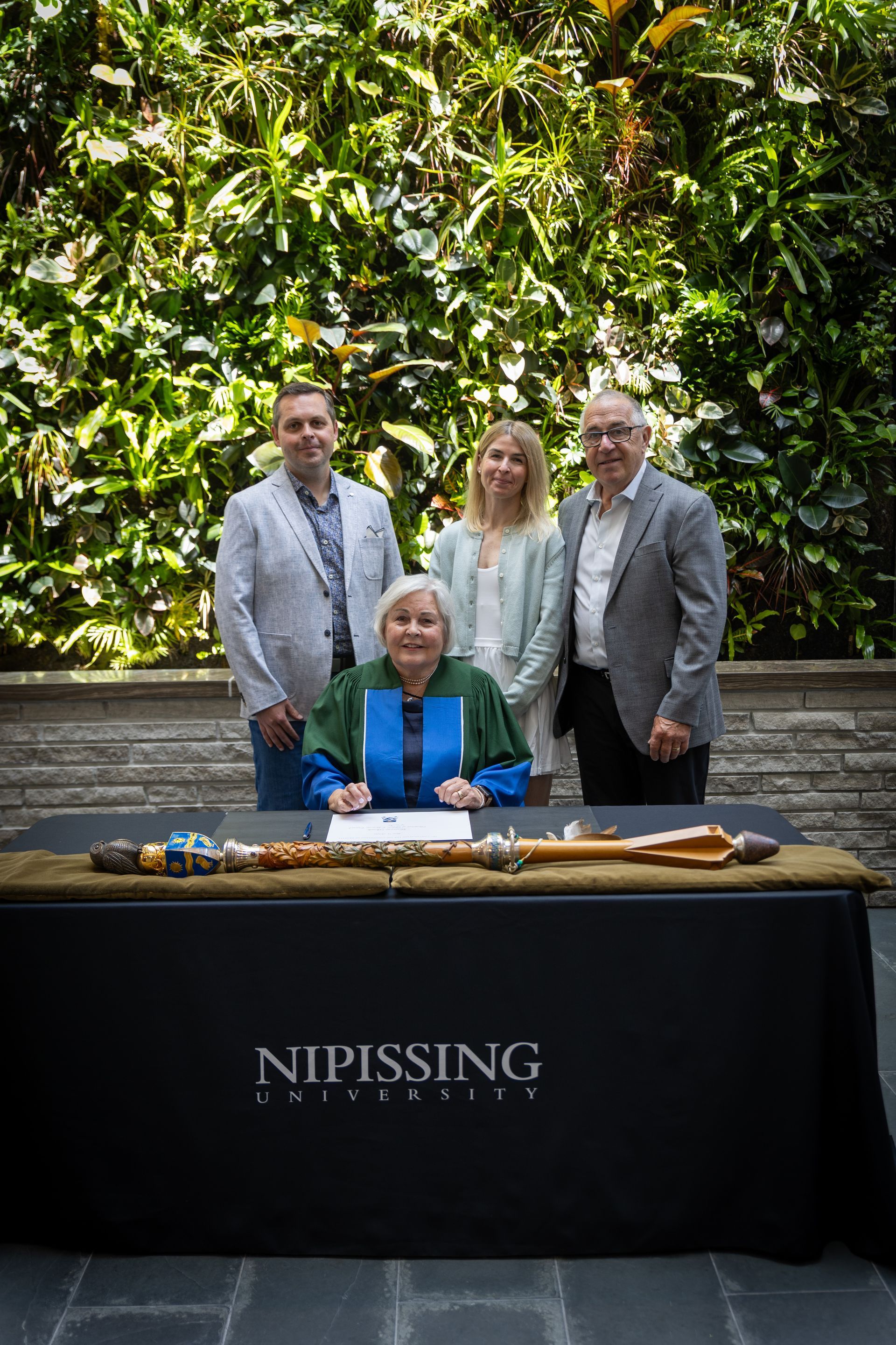 Four people pose behind a signing table with a “Nipissing” cloth in front of a leafy green wall.
