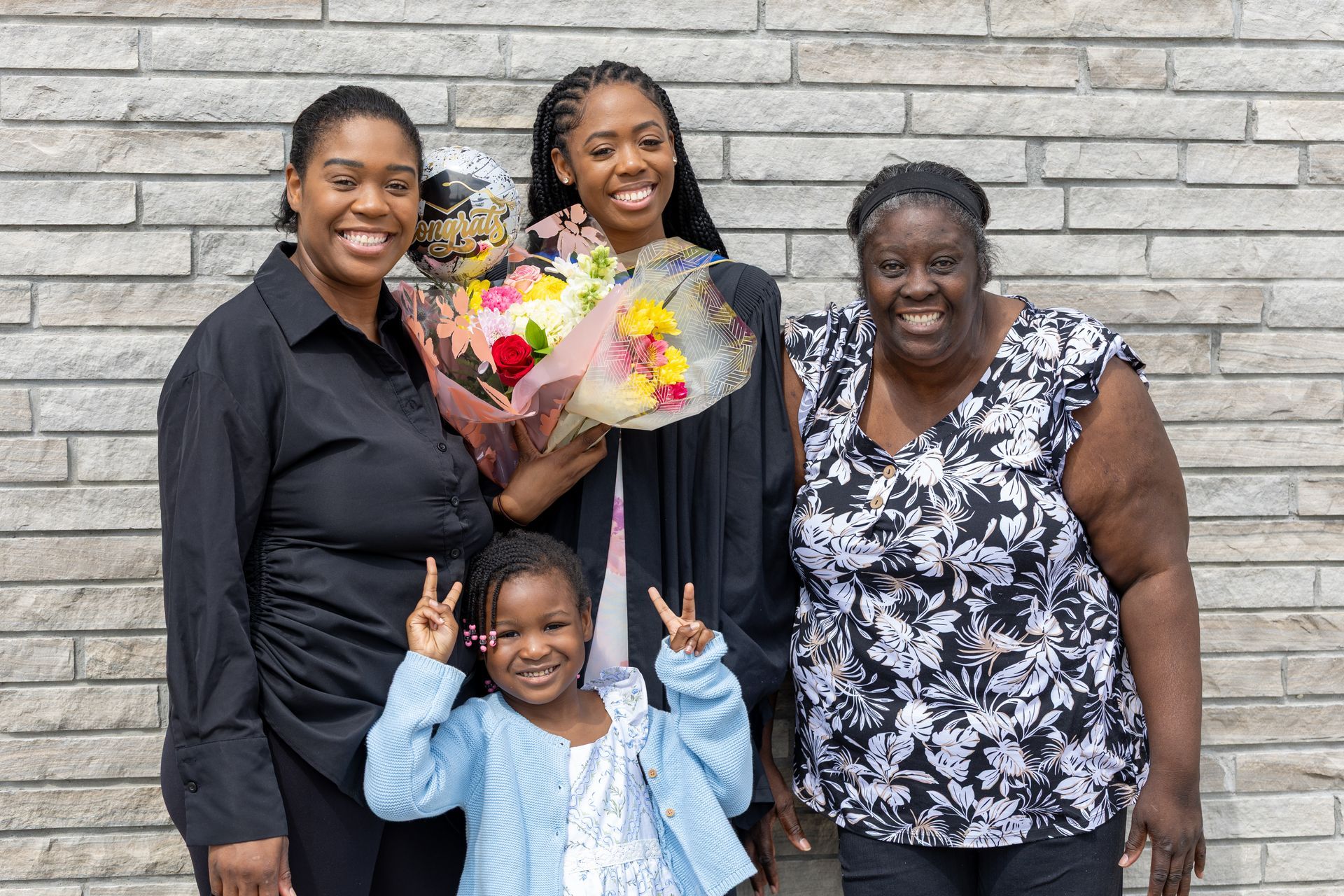 Four people smiling in graduation attire, holding flowers in front of a brick wall
