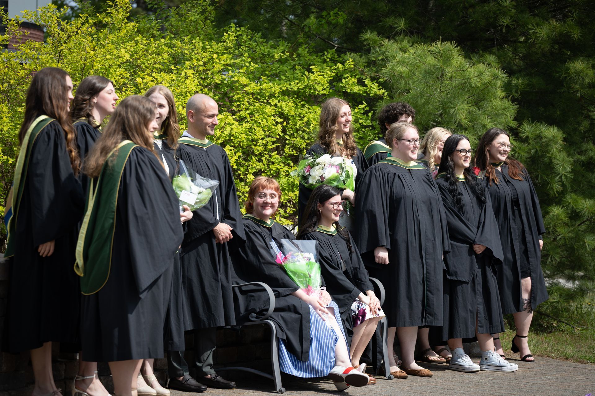 Graduates in black gowns stand outdoors holding bouquets during a ceremony