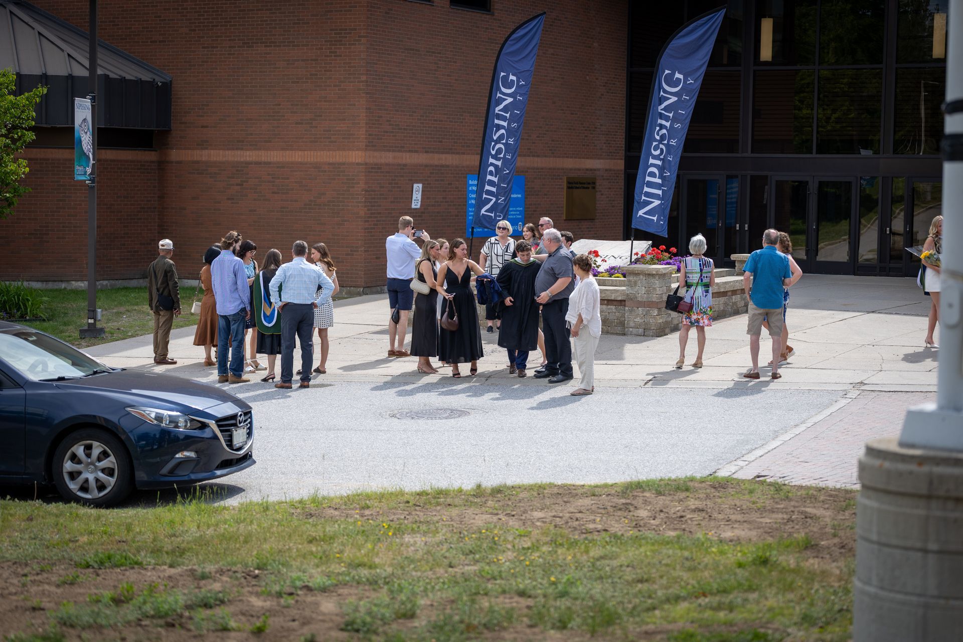 People gathered outside Nipissing University near blue flags, with a parked car in the foreground.