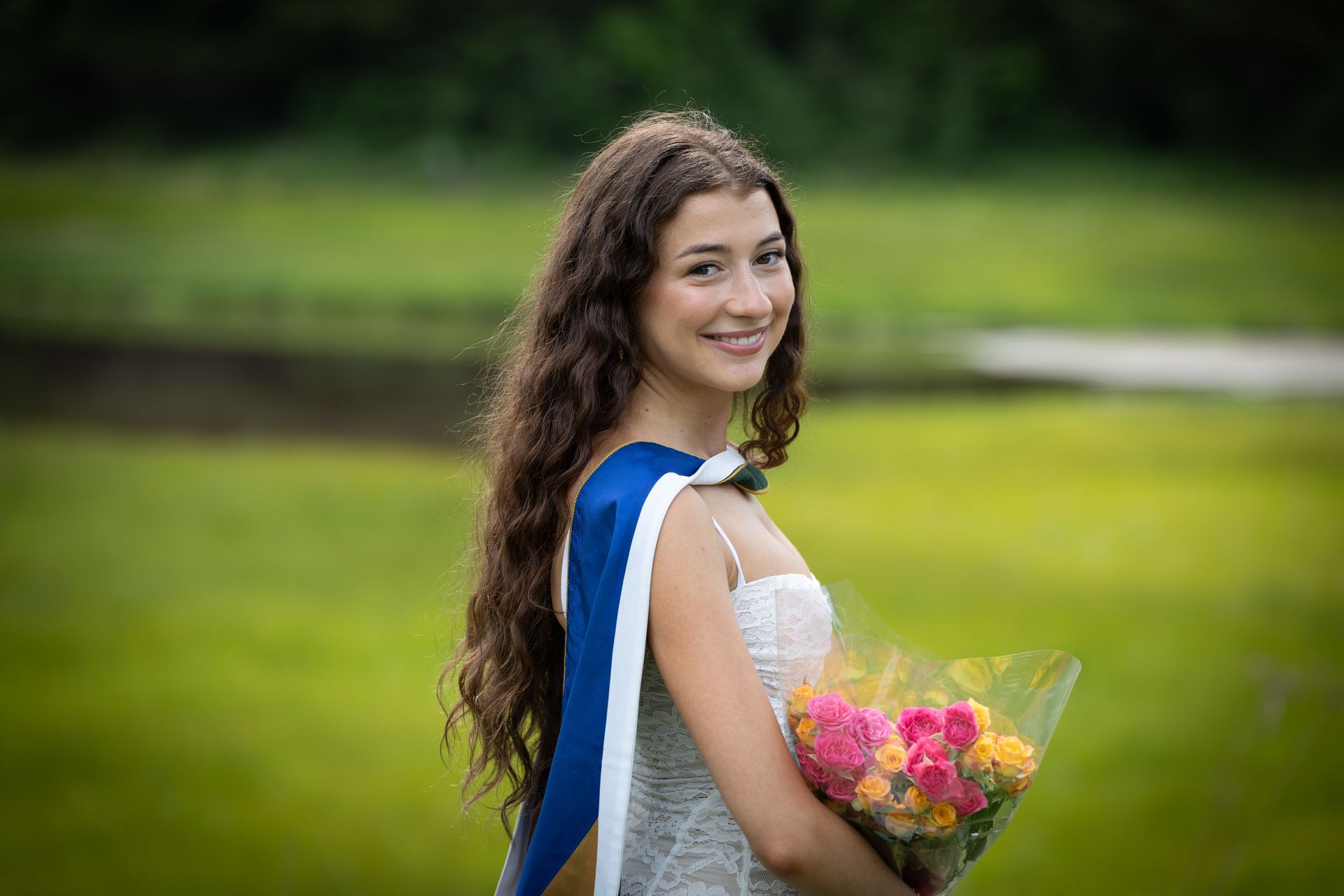 Smiling graduate in blue stole holding a bouquet outdoors on a green lawn