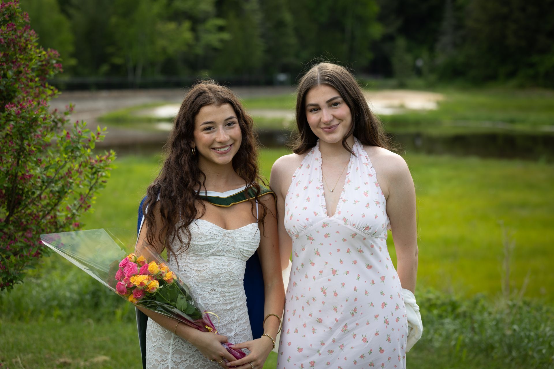 Two women in dresses holding a bouquet outdoors on a green lawn, smiling at the camera.