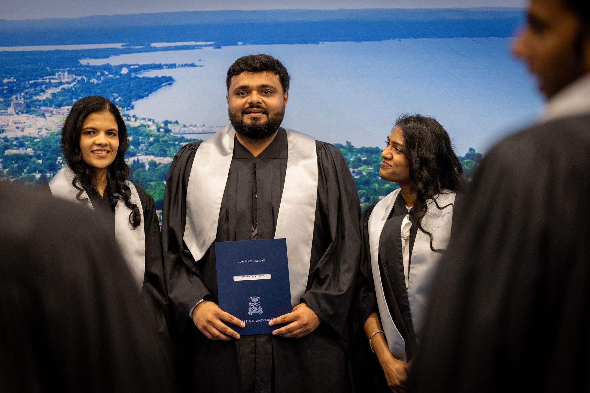 Three graduates in black robes pose before a blue scenic backdrop.