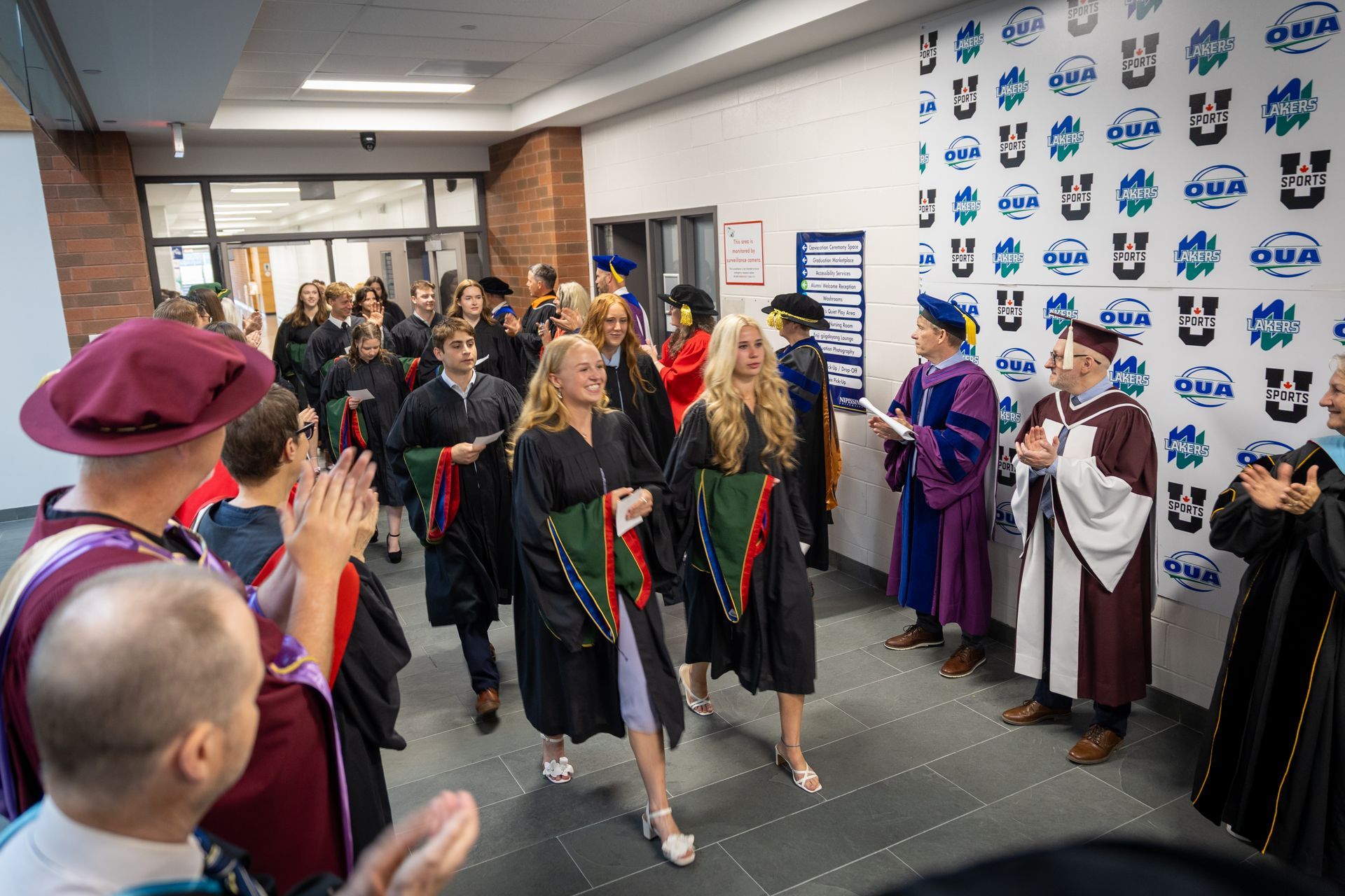 People in graduation gowns and caps applaud in a lobby with a banner-covered wall.
