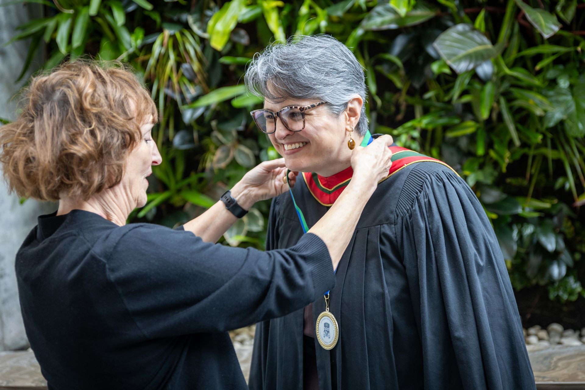 Two people adjusting a black graduation gown and red collar, smiling in front of greenery