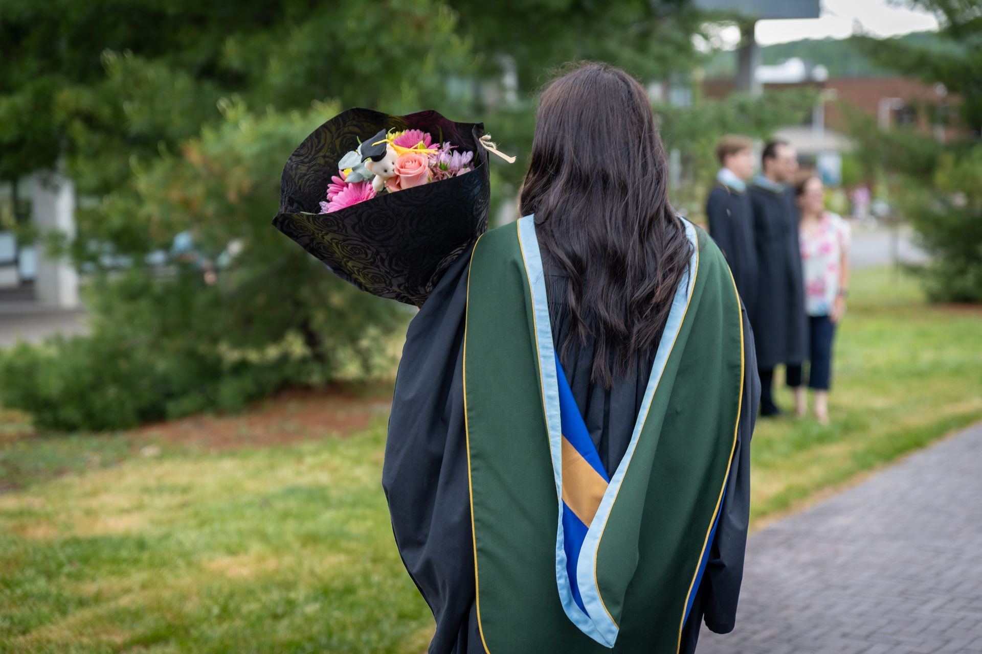 Graduate in gown walking outdoors, holding bouquet, with another graduate ahead on a path