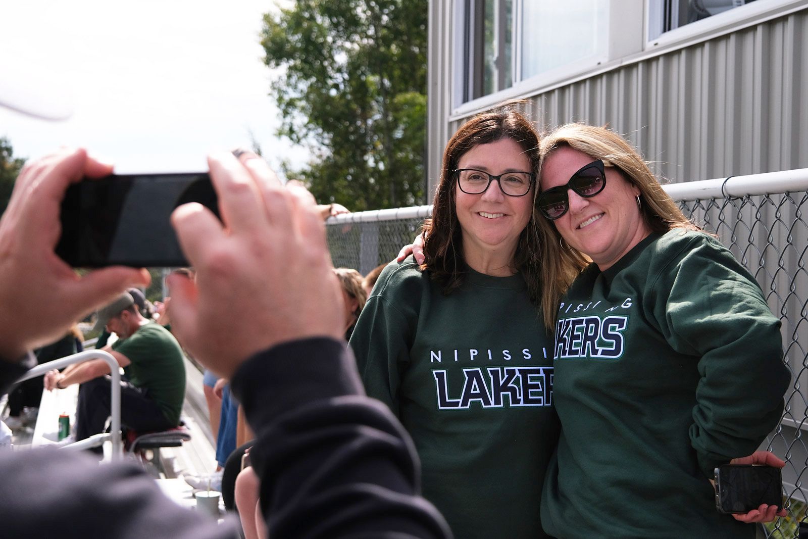 Two people in green hoodies smile for a photo outdoors while someone in the foreground takes it on a phone.