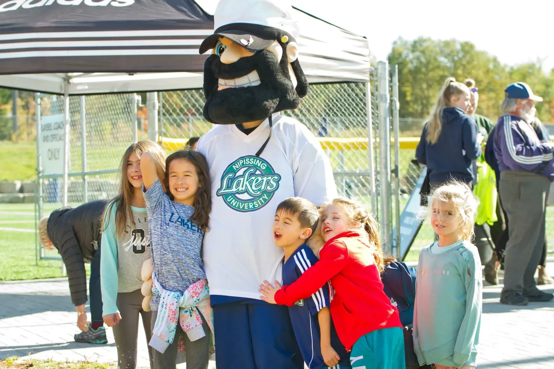Mascot posing with children beside a black canopy tent on a sunny day