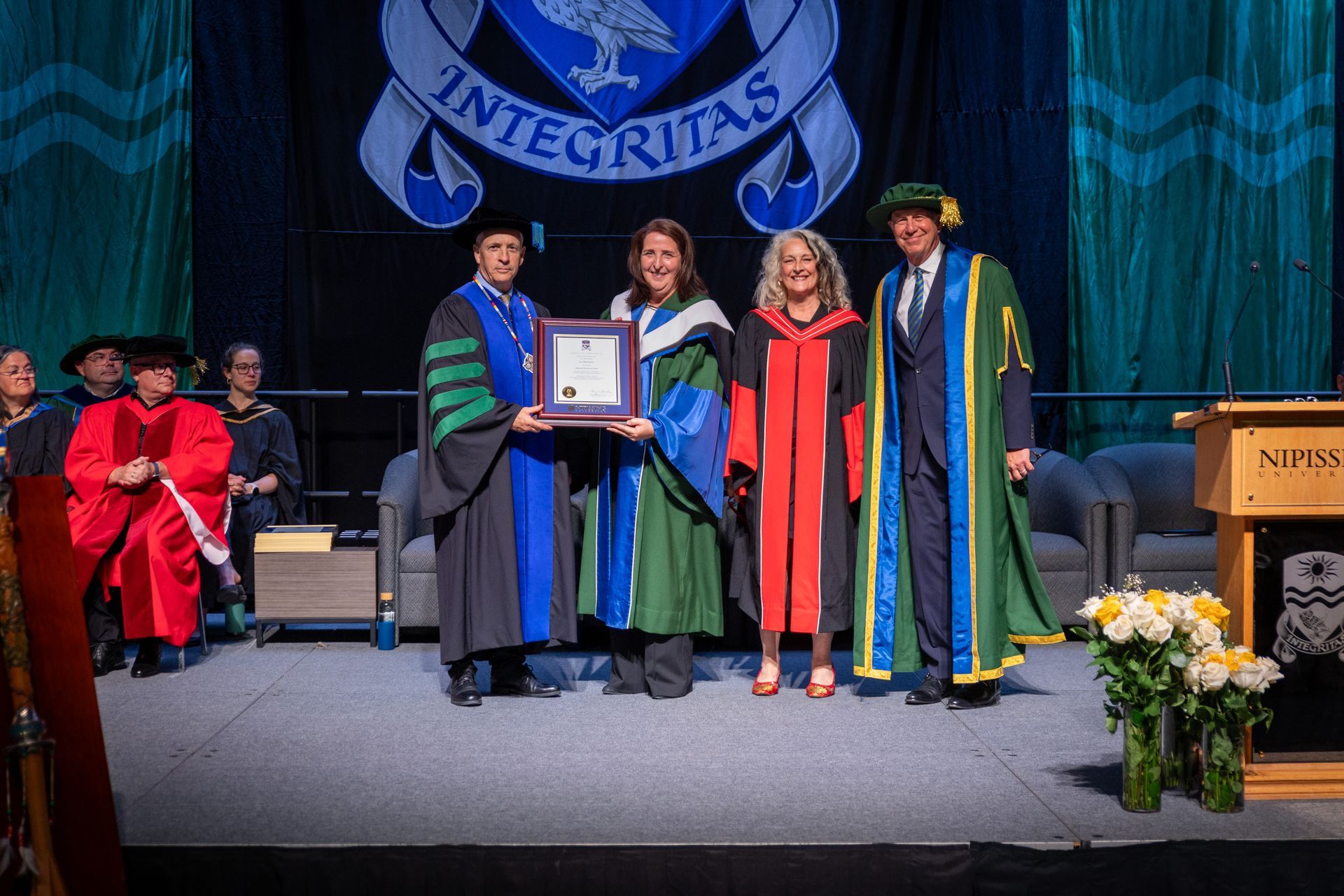 People onstage at a Convocation ceremony, one holding a framed certificate beside faculty in regalia