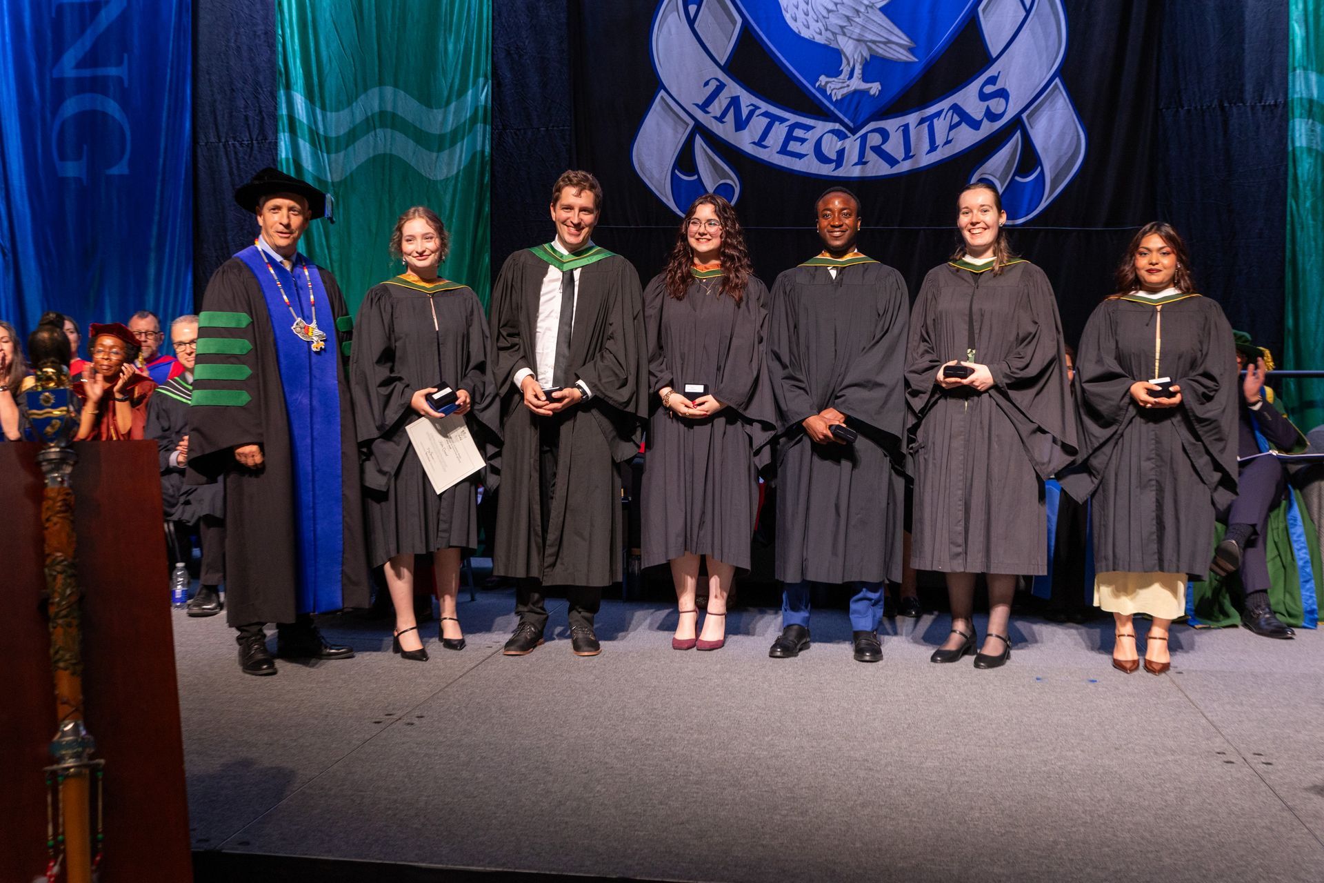 Graduates in gowns pose onstage with faculty before a blue university seal backdrop
