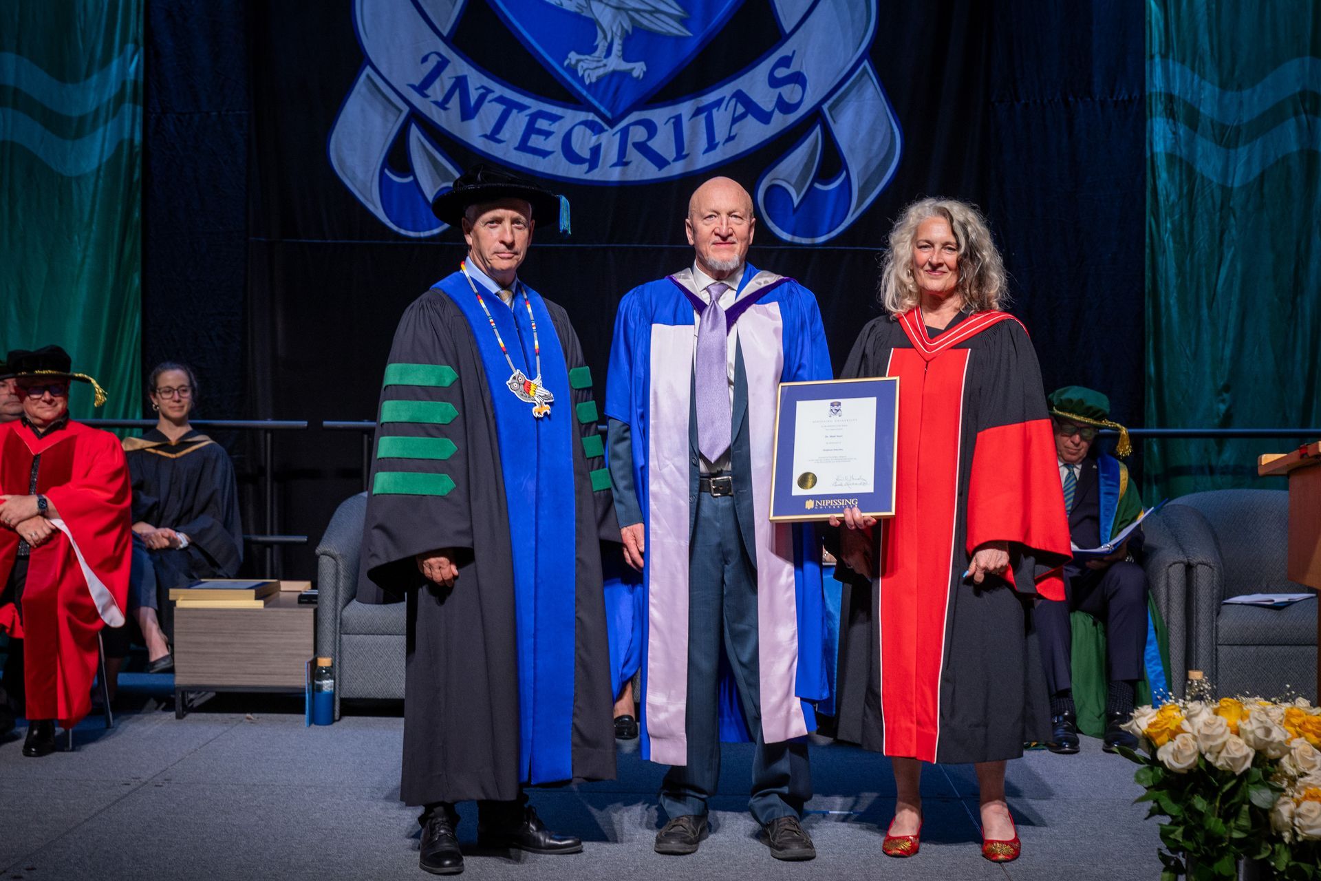 Three people onstage at a graduation ceremony, in academic robes holding a certificate.