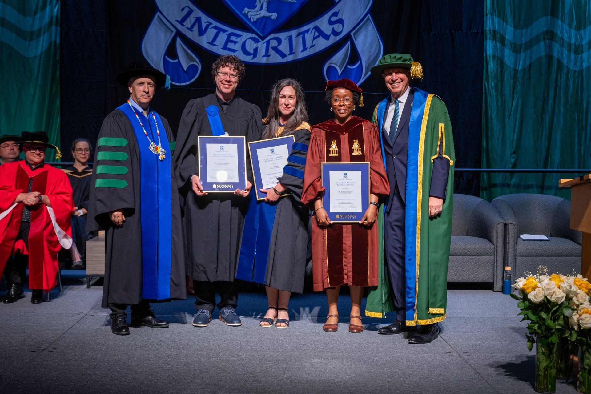 Five people in graduation regalia pose onstage with framed awards in front of a university backdrop