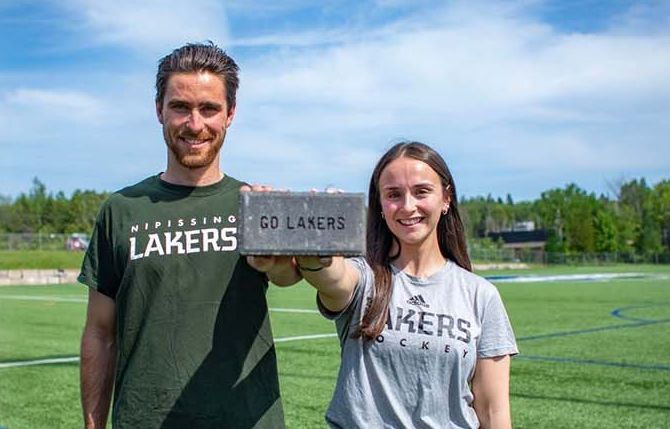 Two people on a field hold a “Go Lakers” brick, wearing Lakers shirts and smiling in sunlight.