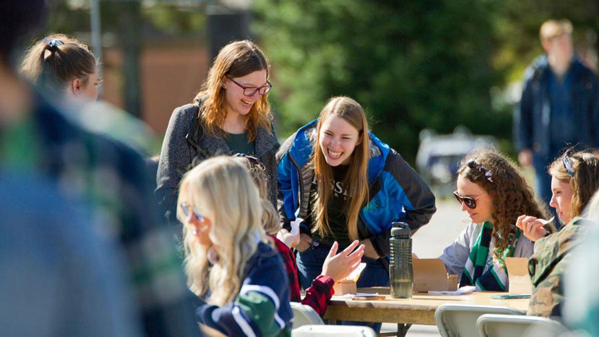 People chatting outdoors at a picnic table on a sunny campus lawn