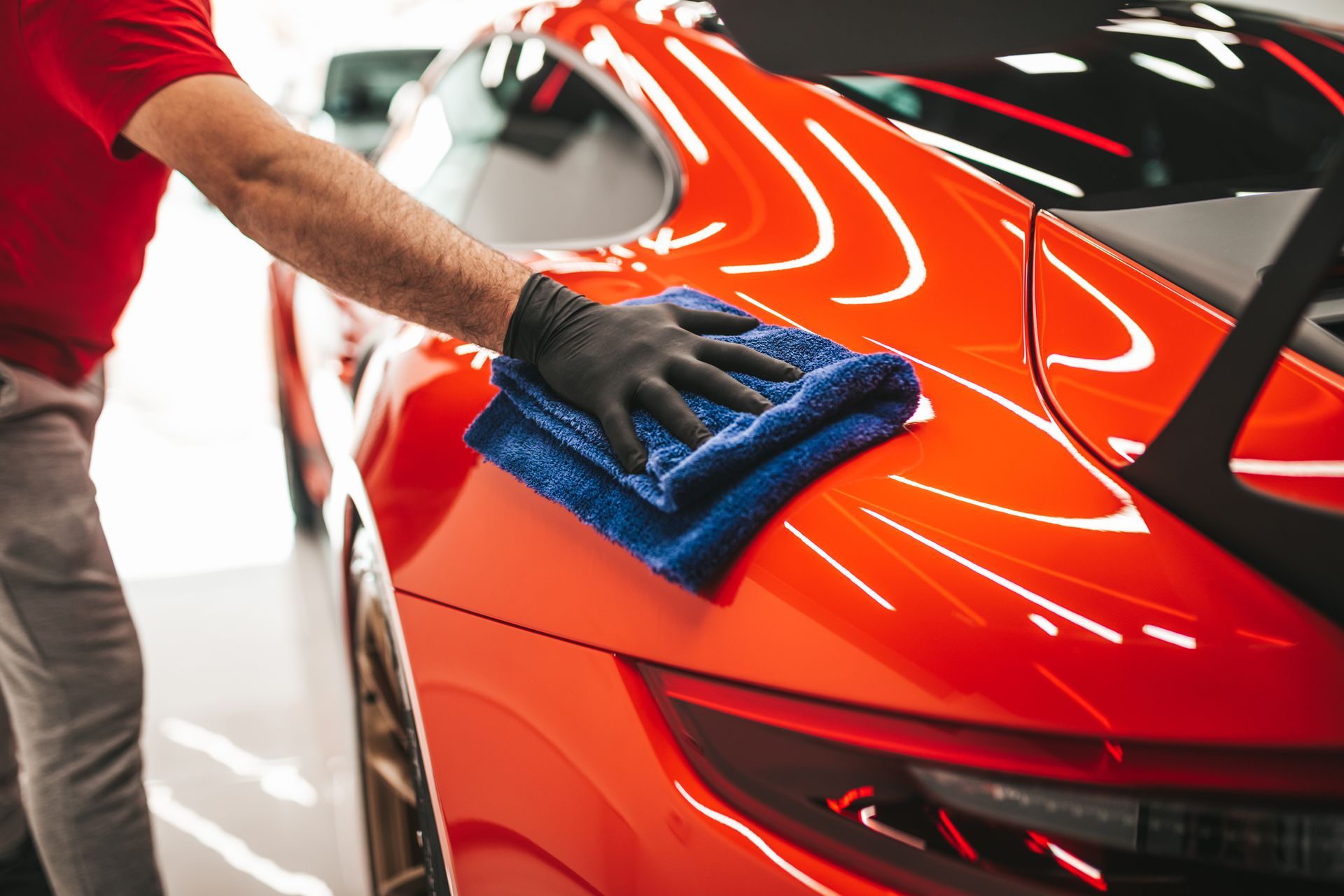 Person in black glove wiping a vibrant red sports car with a blue microfiber towel.