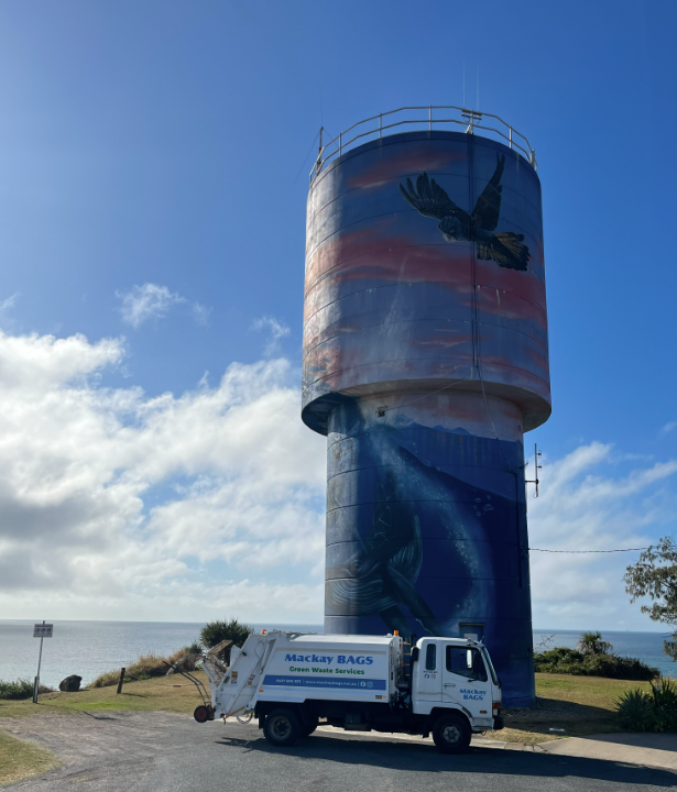 A green garbage truck is parked in a field with trees in the background — Mackay Bags in East Mackay, QLD