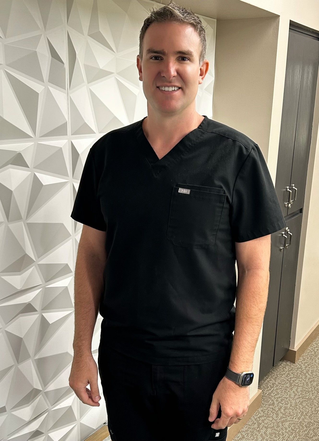 A man in a black scrub top is standing in front of a white wall.