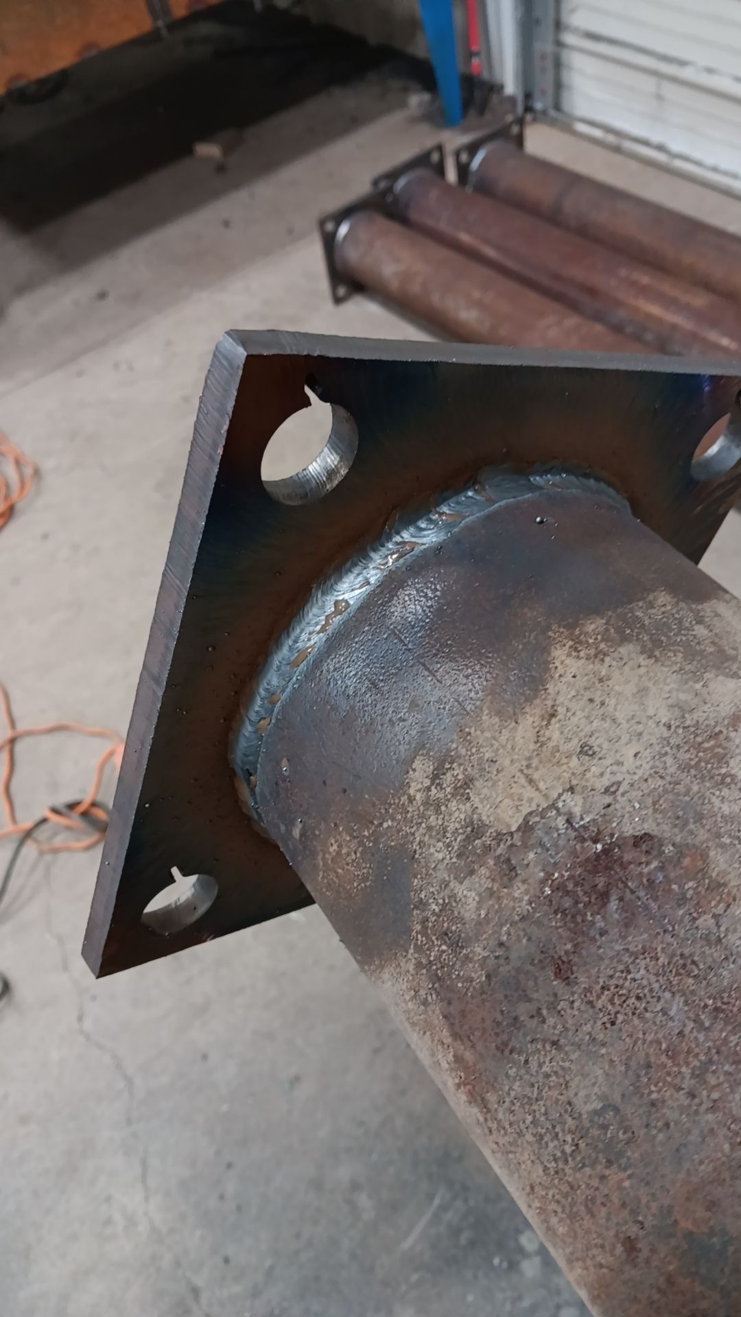 Rusty metal pipe bolted to a square flange on a concrete floor in an industrial setting