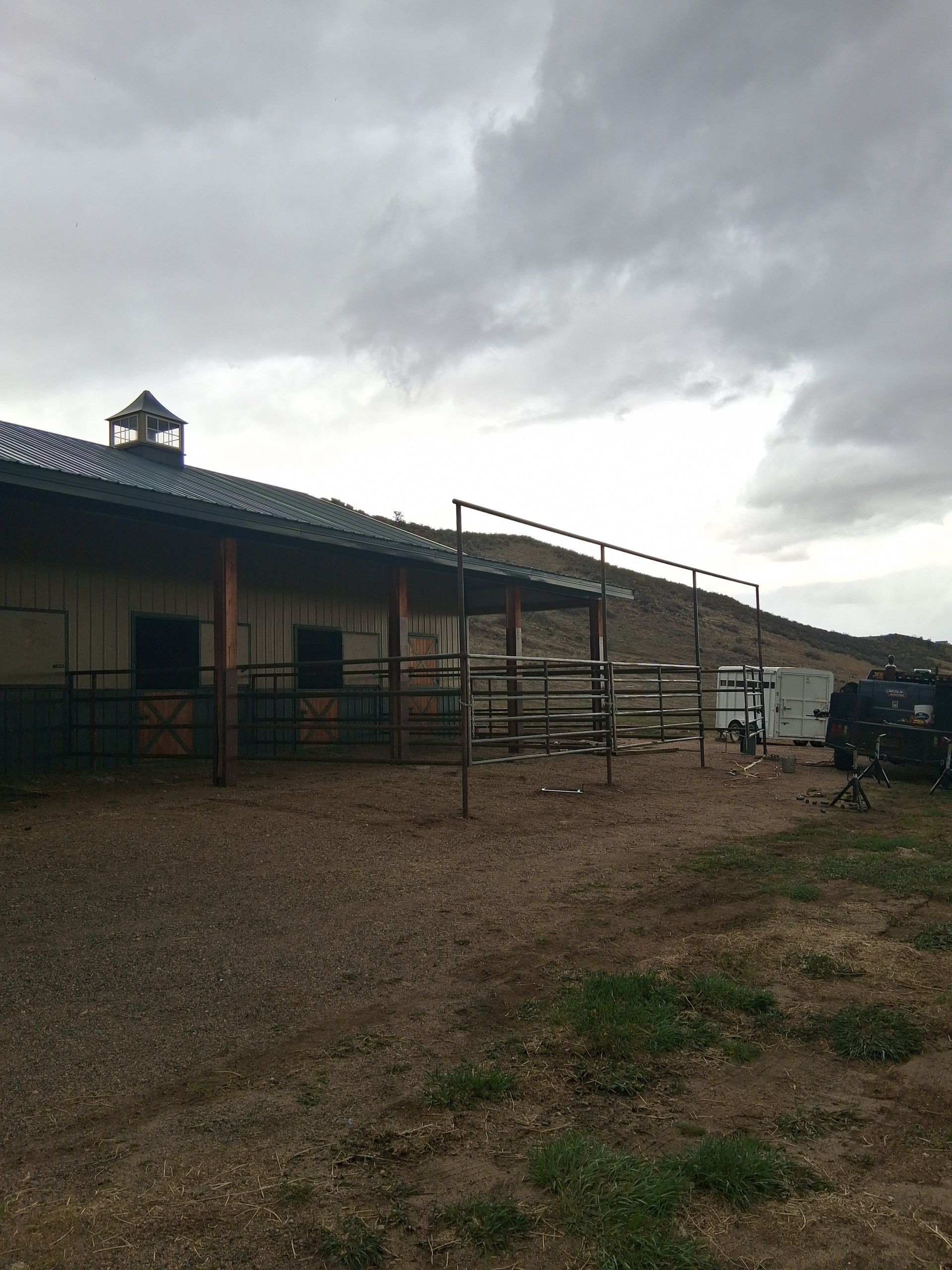 Cloudy ranch yard with a long metal-roofed barn, gravel ground, and fencing under dark storm clouds